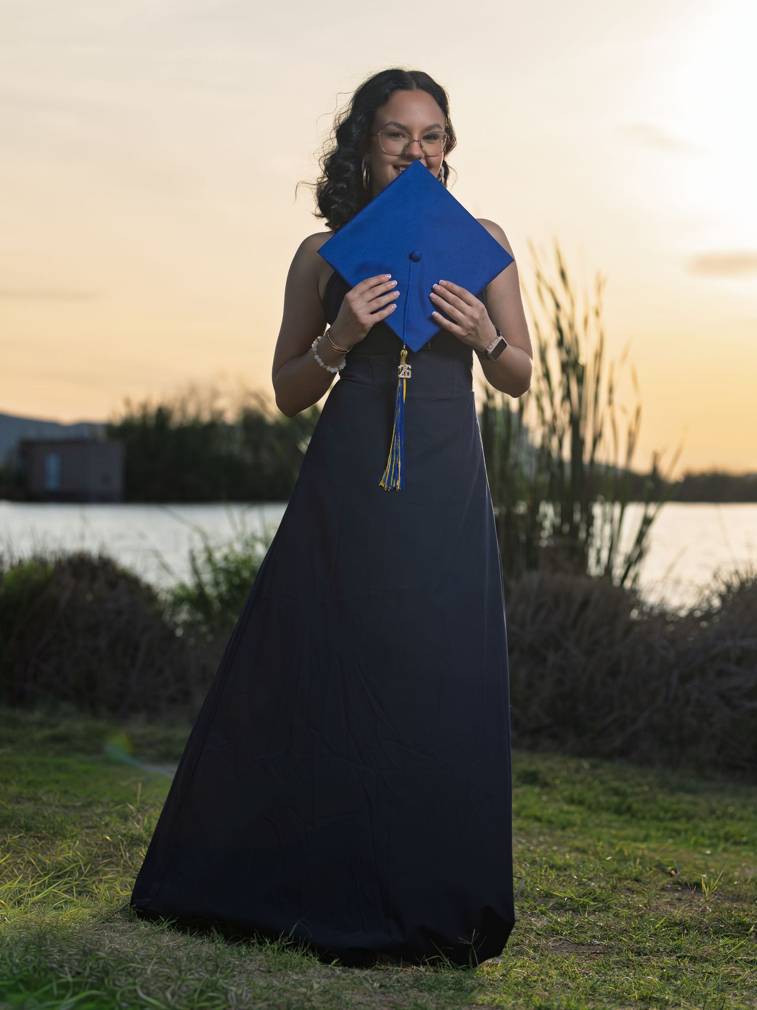 Graduate in navy blue dress holds a blue diploma folder against sunset sky by waterfront during outdoor graduation photoshoot.