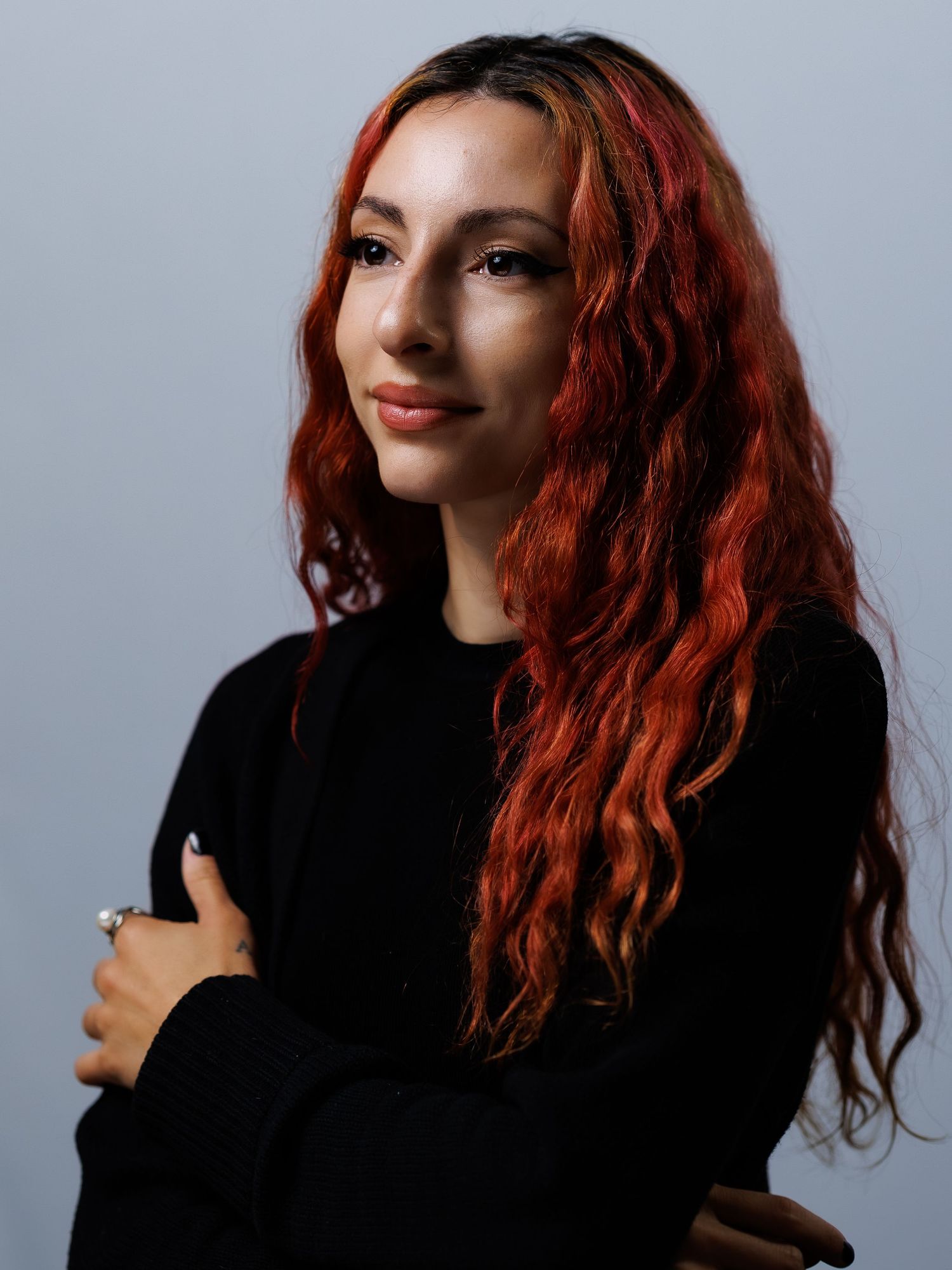 A person with vibrant red curly hair wearing a black sweater poses against a gray background in a photo series.