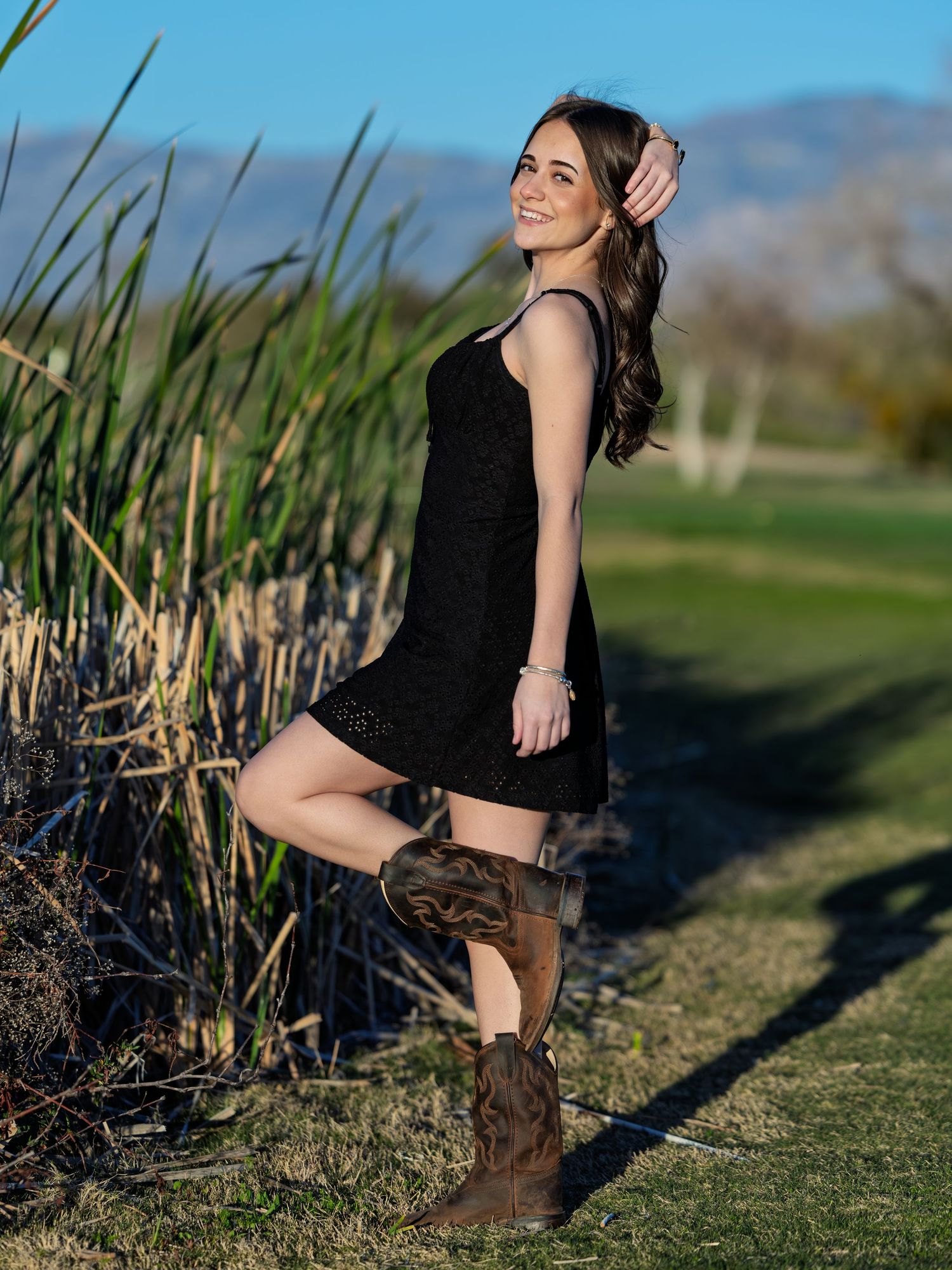 A dancer in a black outfit and boots strikes graceful poses near tall grass at sunset in a rural outdoor setting.