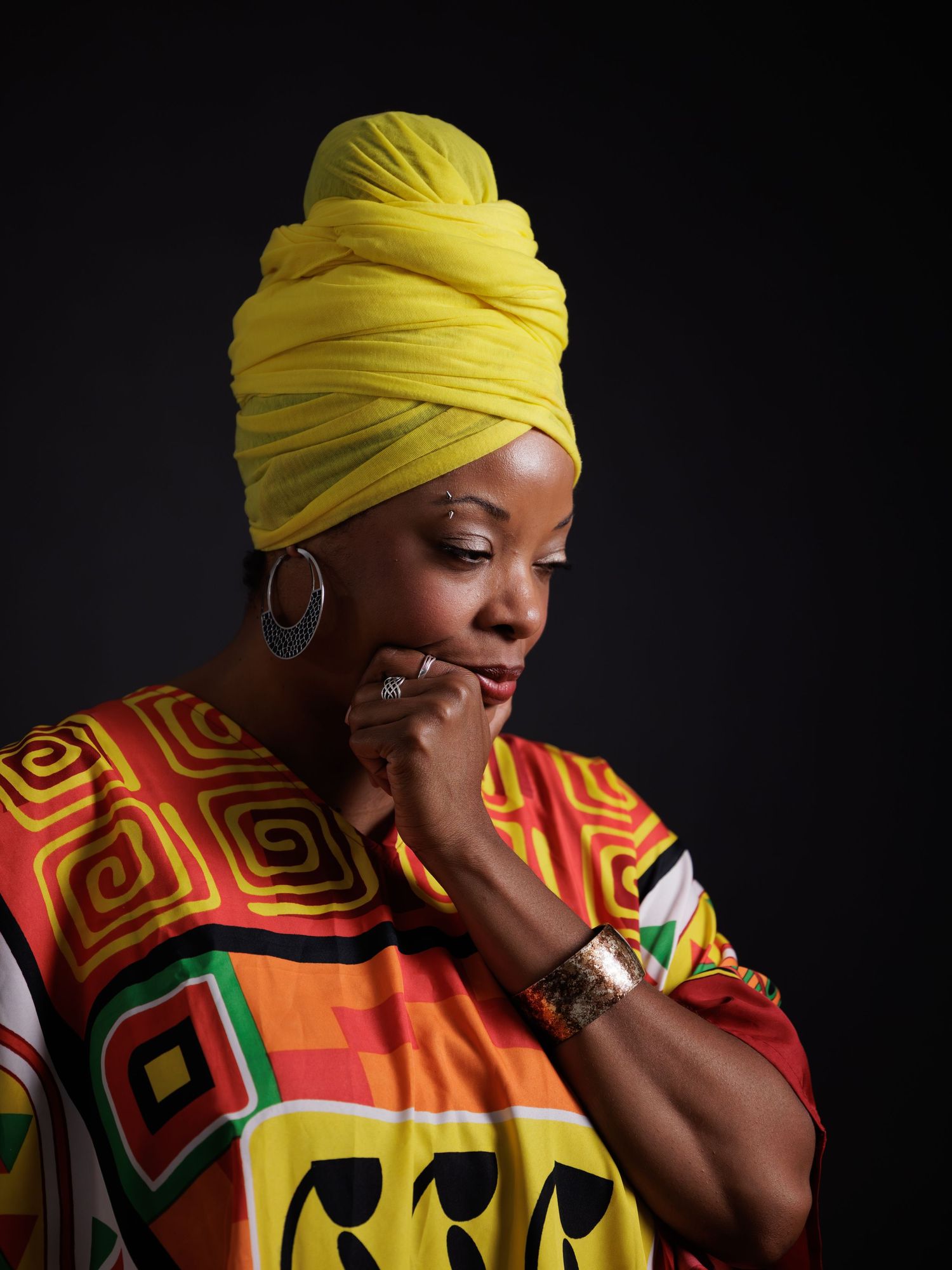 A figure in vibrant African print clothing and yellow headwrap poses expressively against a dark background.