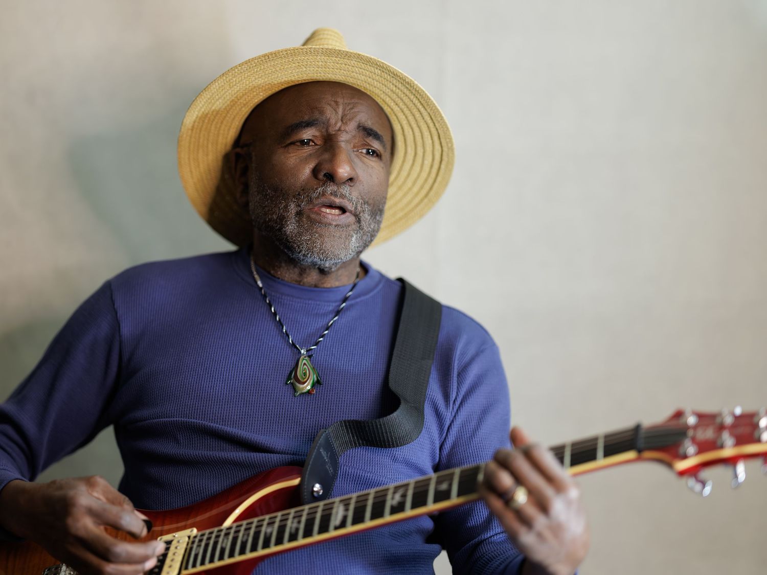 A musician in a blue shirt and straw hat plays an acoustic guitar while seated against a light colored background.