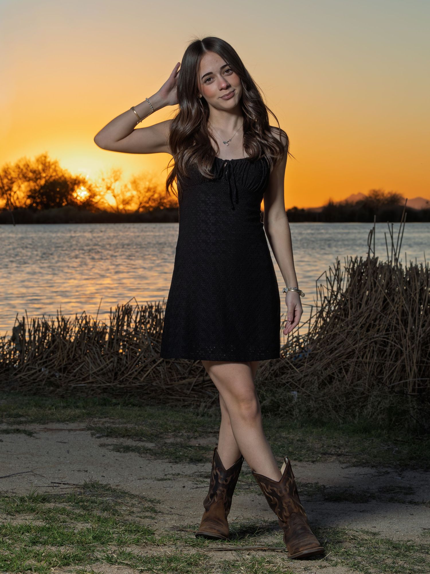 Woman in black dress and cowboy boots poses against stunning sunset by lake with tall grass silhouettes.