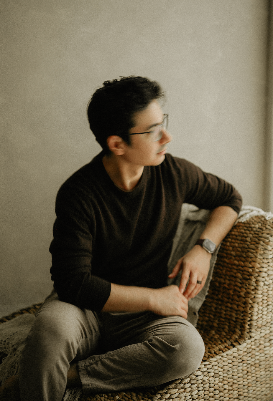 Man with glasses and a dark sweater sits on a woven wicker chair, looking to the side in soft natural light.