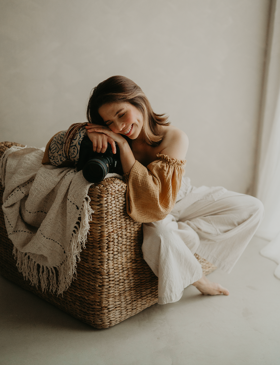 Smiling woman in a mustard off-shoulder top and cream pants rests her head on a camera atop a woven wicker chair with a fringed throw.