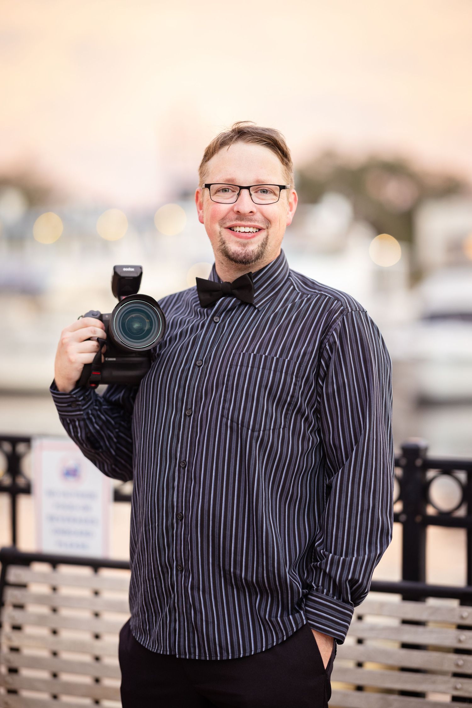Professional photographer in striped button-down shirt poses outdoors with camera equipment against marina backdrop.