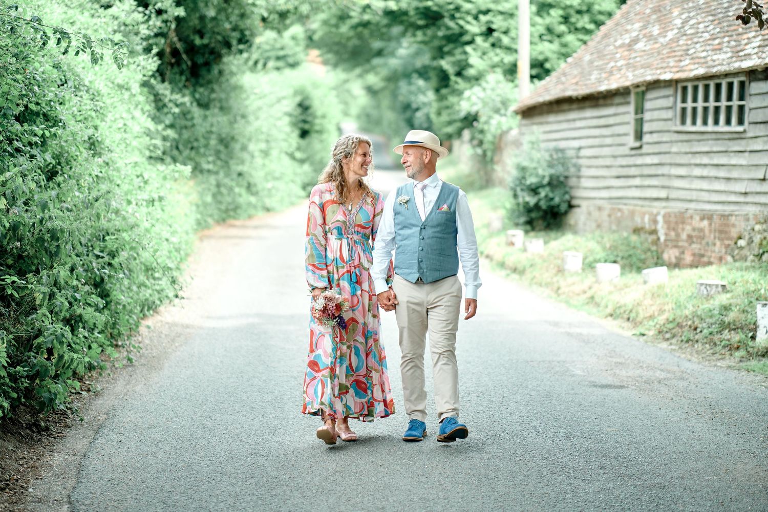 A couple walking together on a rural country road wearing summer attire with a wooden building in the background.