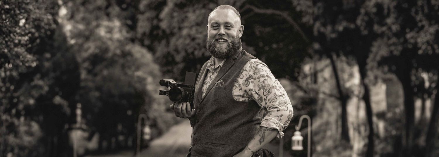 A photographer with camera gear smiles while capturing outdoor photos in a park setting.