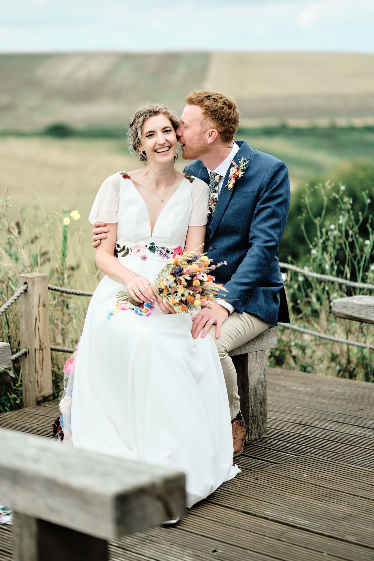 Newlywed couple shares intimate moment on rustic wooden boardwalk surrounded by wildflower meadow at sunset.