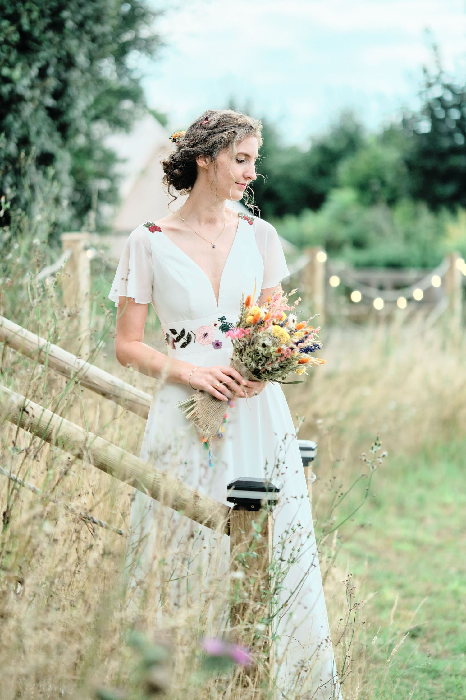 A person in a white lace dress holds a colorful bouquet while walking through a rustic field with wooden fencing.