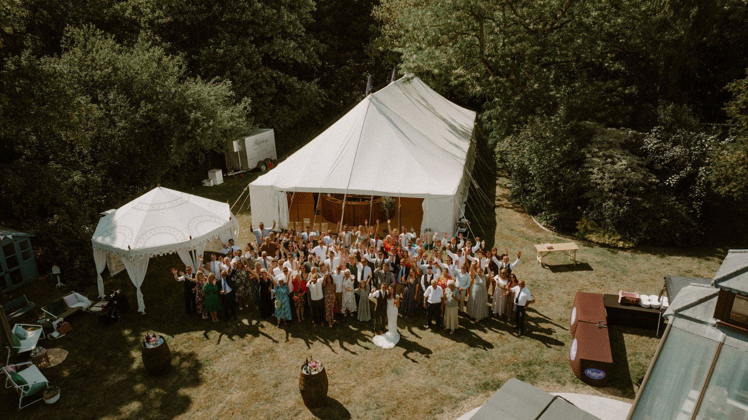 Aerial view of an outdoor wedding reception with white tents and gathered guests.