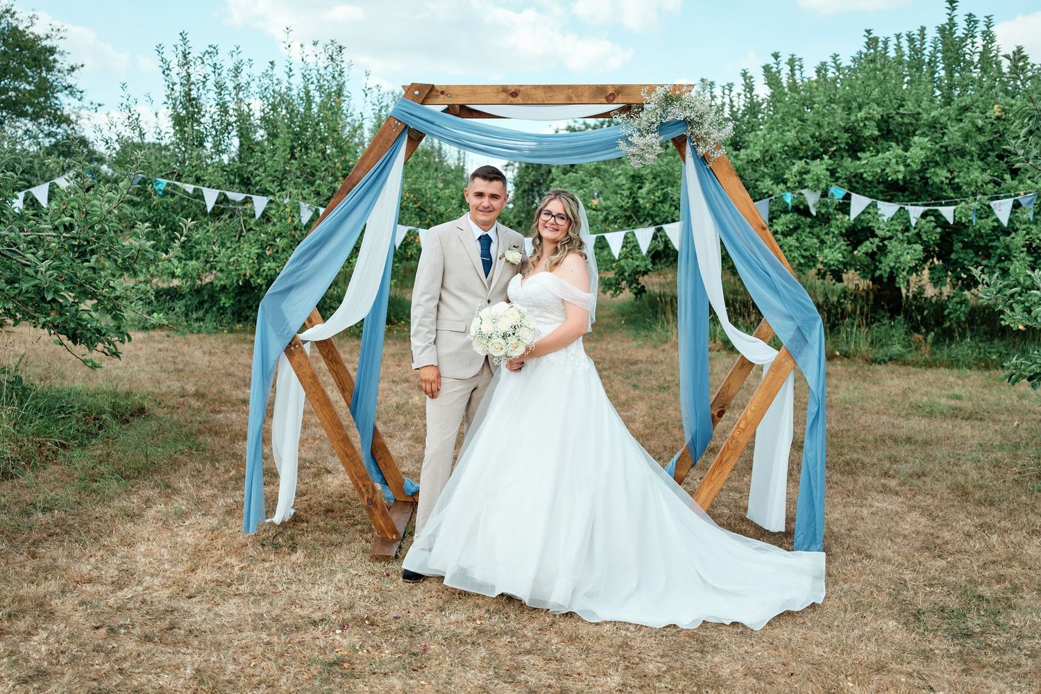 Bride and groom posing under rustic wooden arch with blue draping and bunting at outdoor orchard wedding.
