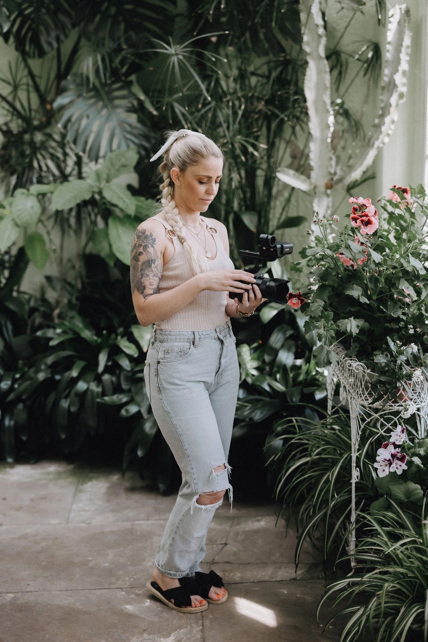 Professional photographer in ripped jeans and floral top checks camera settings in lush greenhouse with blooming flowers.