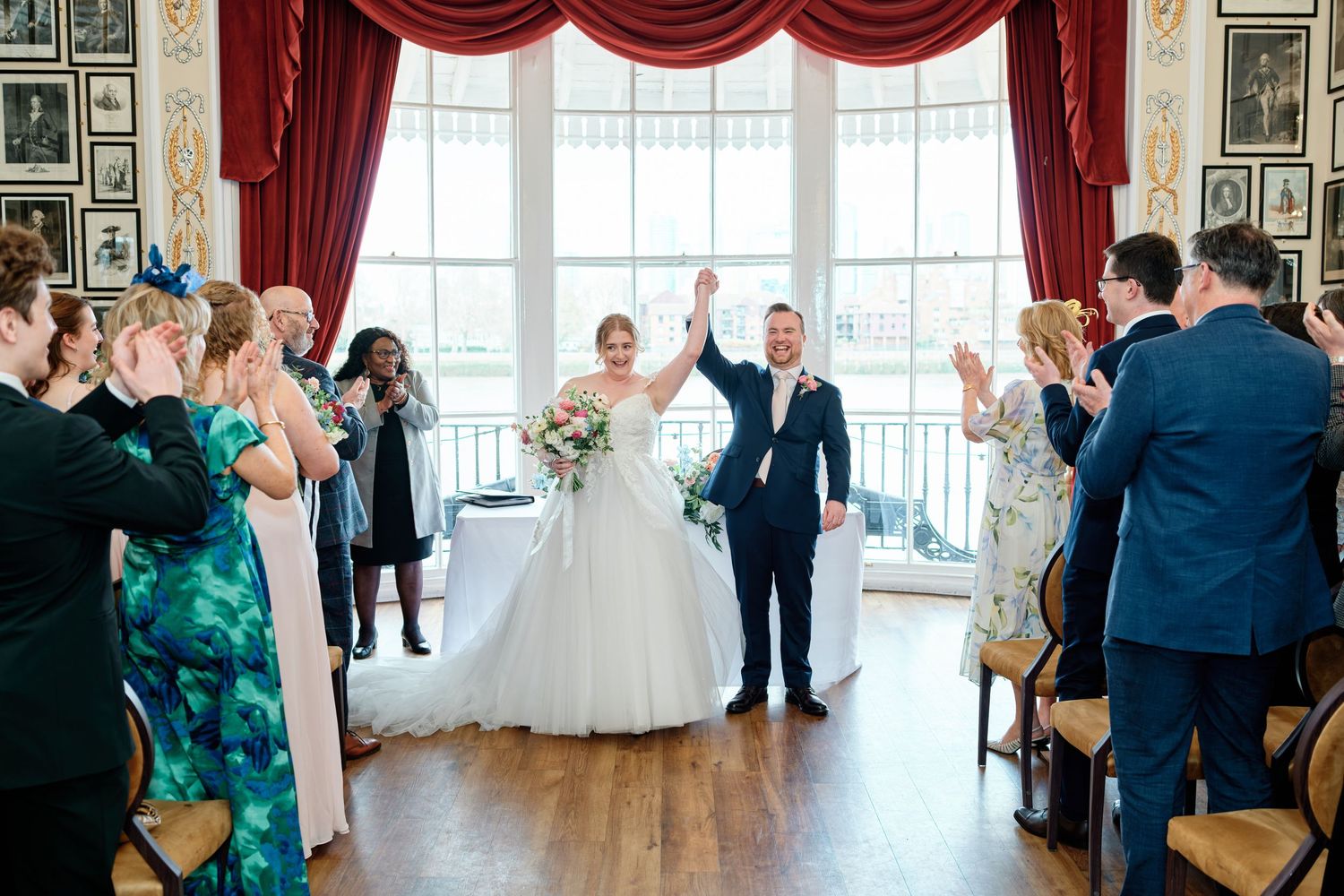 Bride and groom celebrate their wedding ceremony exit, raising hands joyfully as guests applaud at the Trafalgar Tavern, Greenwich