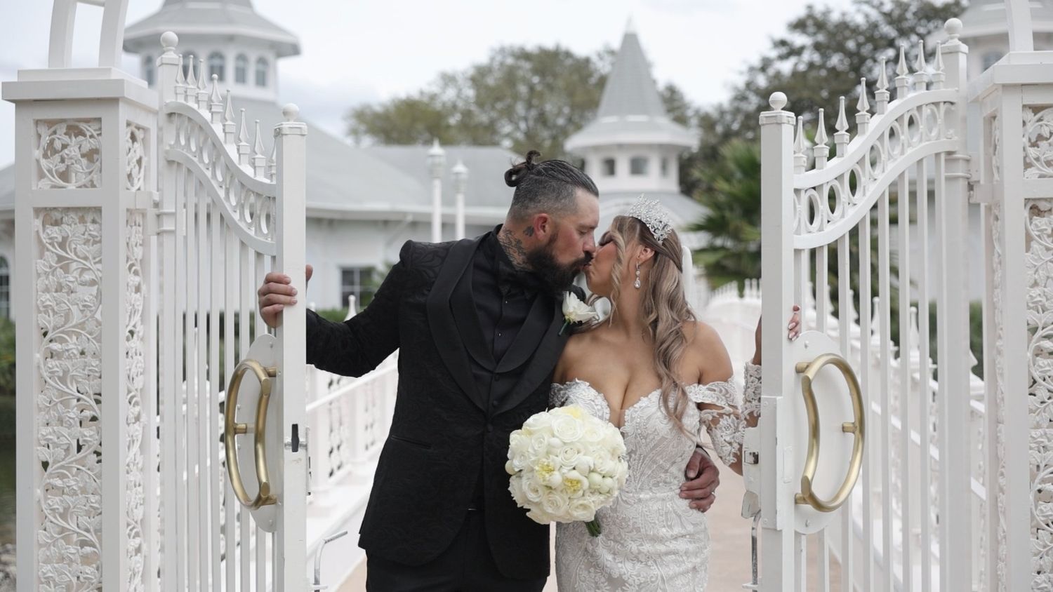 Bride and groom share a romantic kiss at ornate white garden gates on their wedding day.