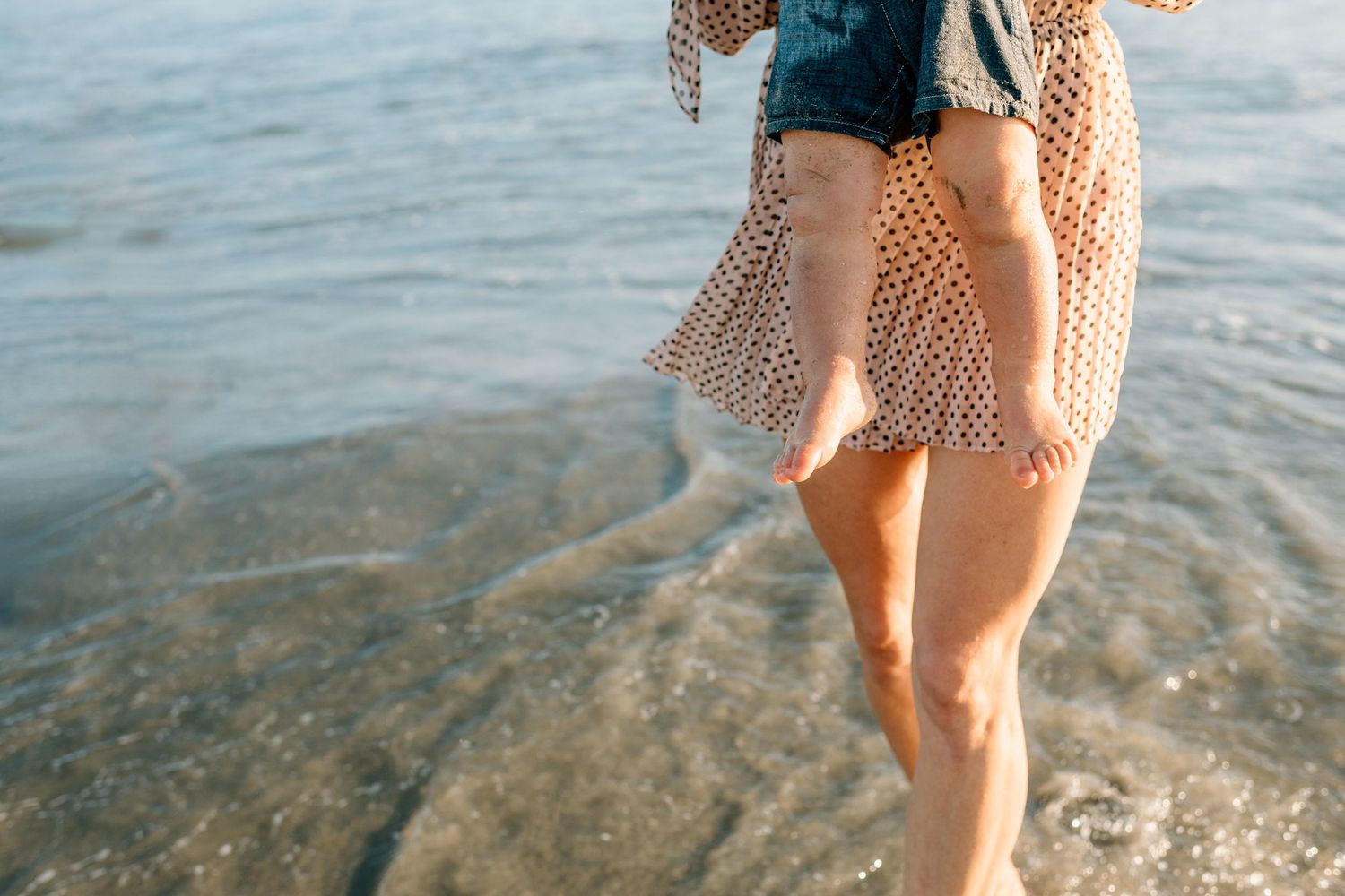 Woman in denim shorts and lace kimono walking barefoot on a sunny beach with gentle waves.