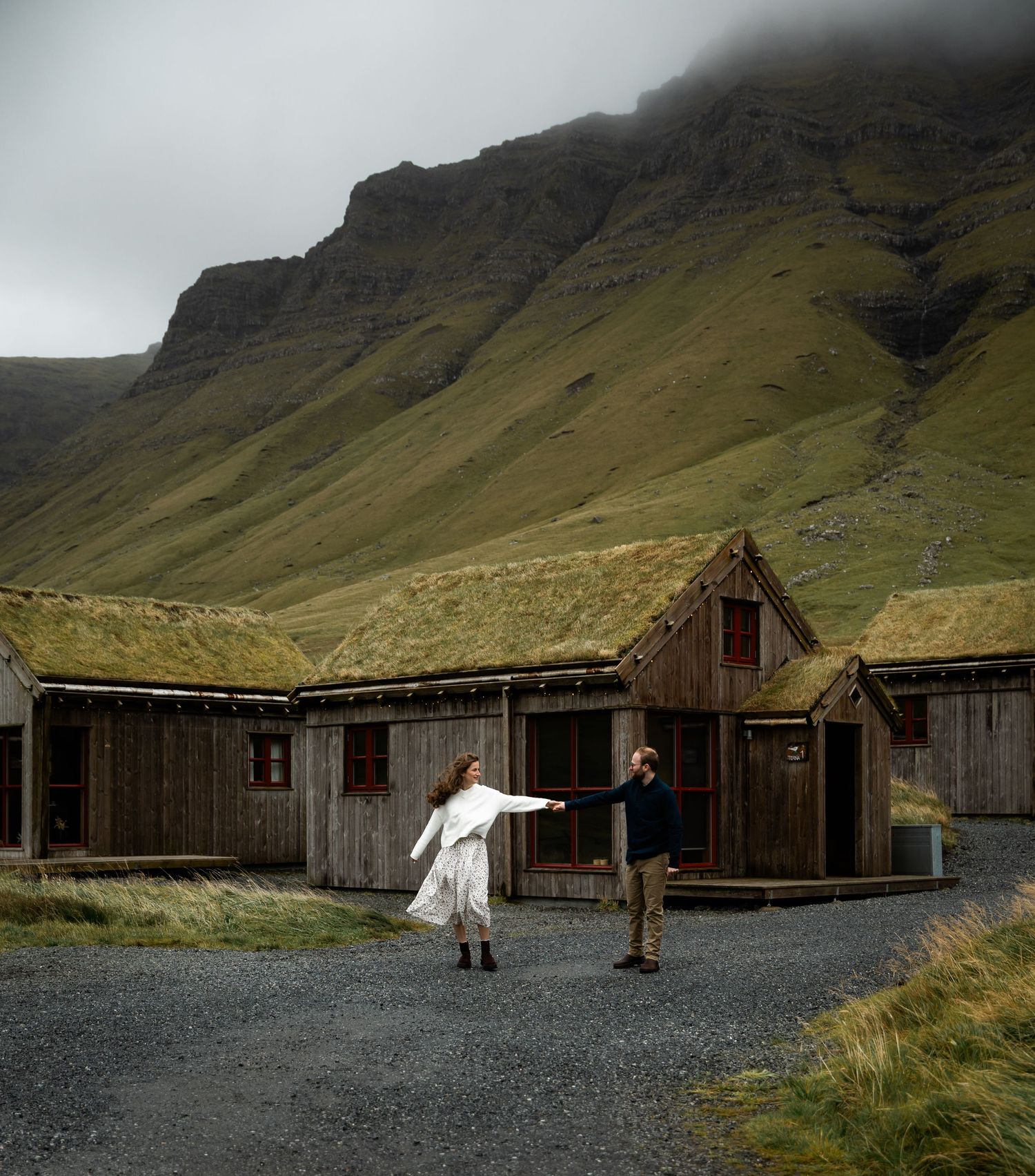 Rustic wooden cabins with grass roofs nestled against dramatic mountain cliffs in a misty Nordic landscape.