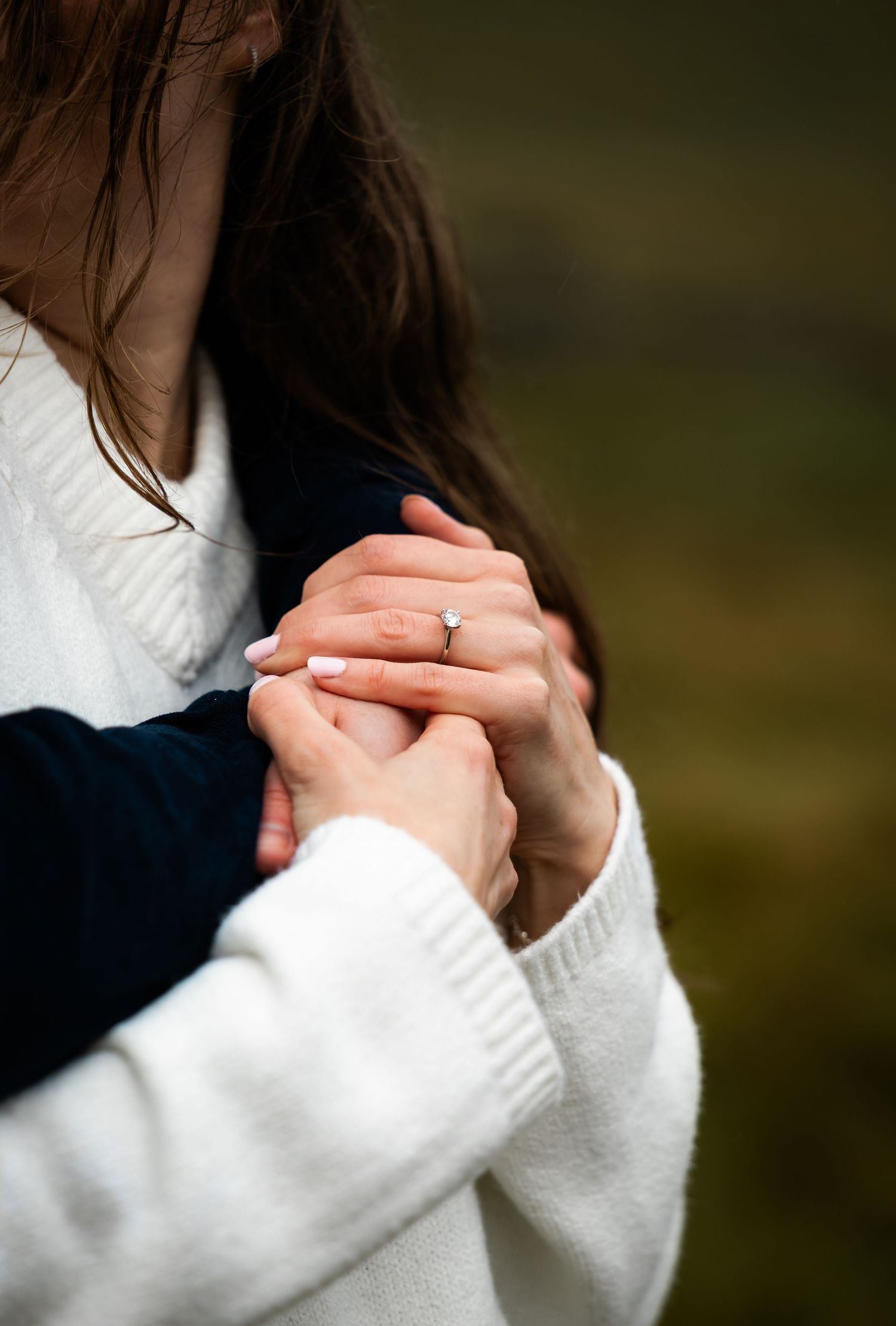 Close-up shot of hands clasped together wearing a white sweater and a delicate engagement ring.