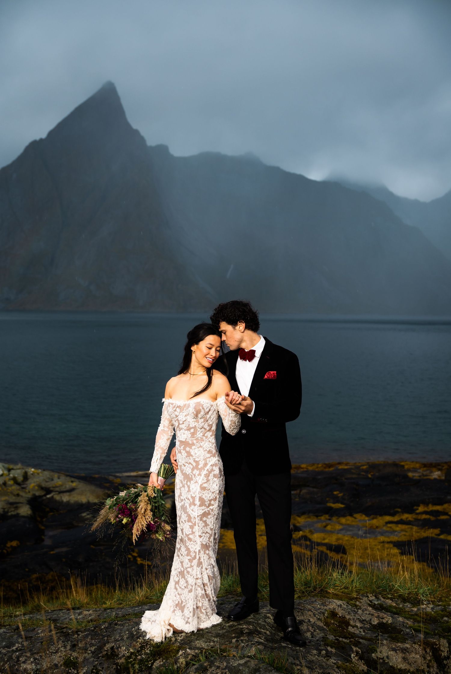 Couple embraces on a dramatic cliffside overlooking misty mountains and a serene lake in moody weather.