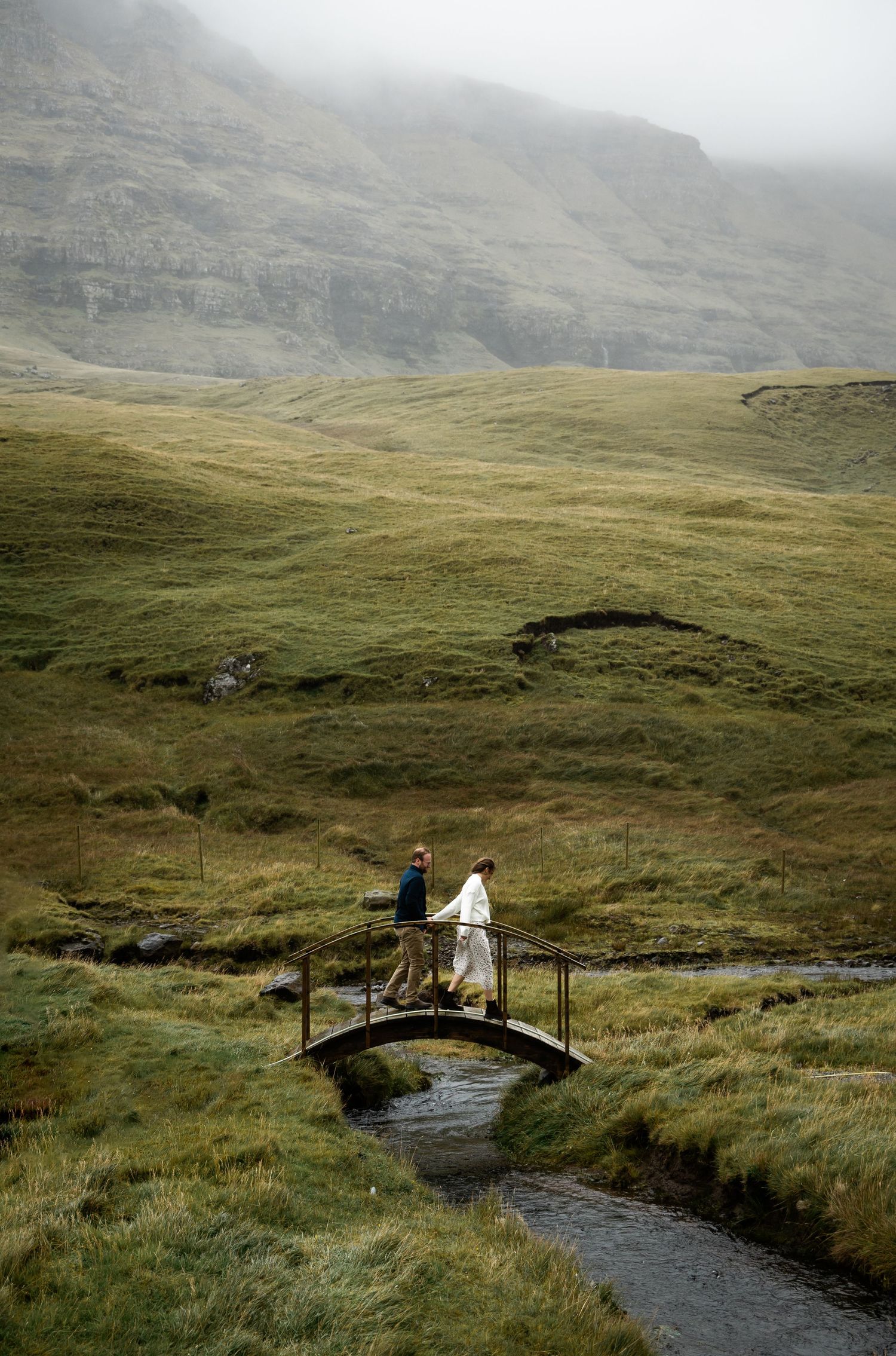 Two figures stand on a small footbridge crossing a stream in misty Scottish highlands with green rolling hills.