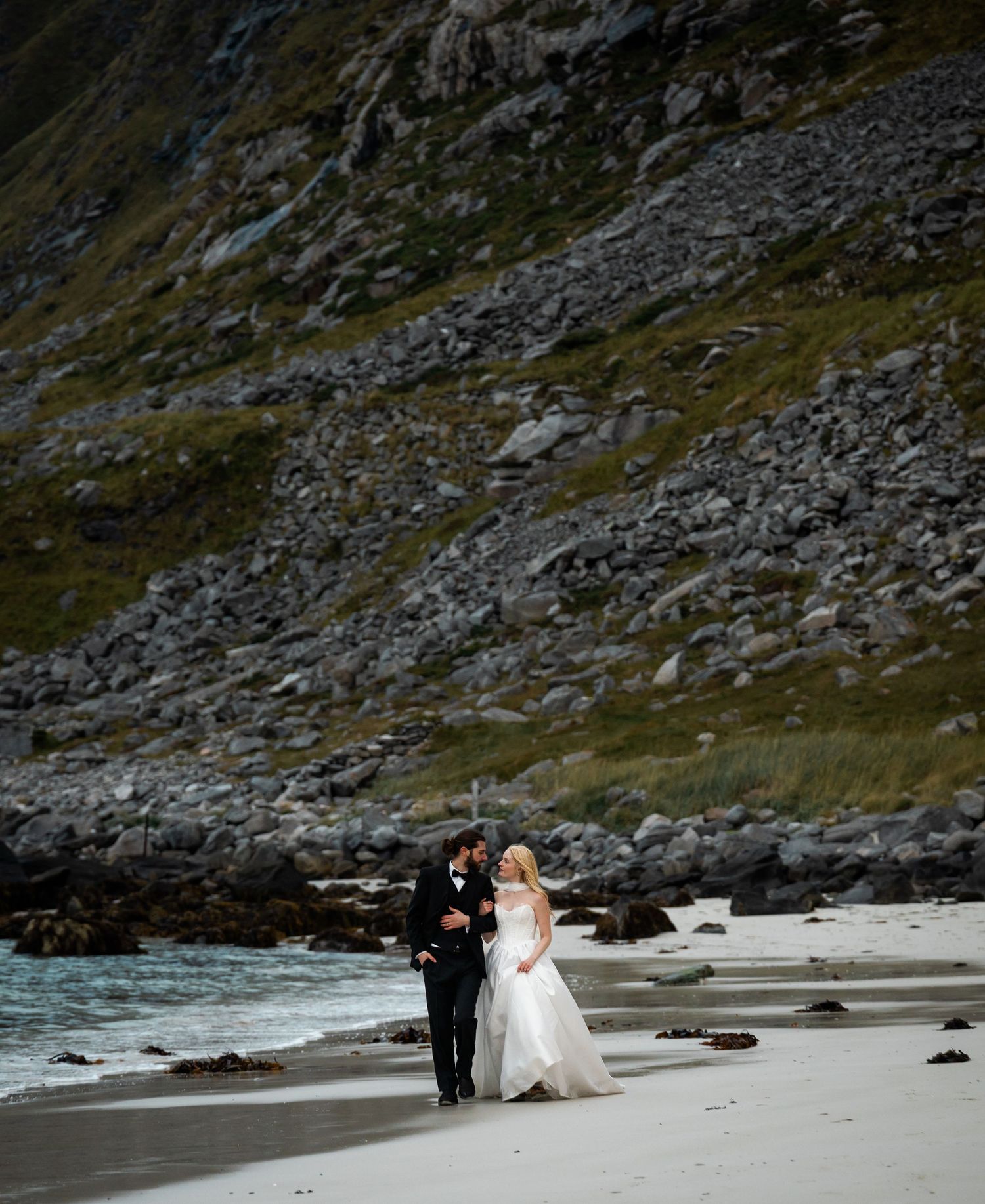 A couple walks along a rocky beach with dramatic mountain cliffs in the background during a wedding photoshoot.