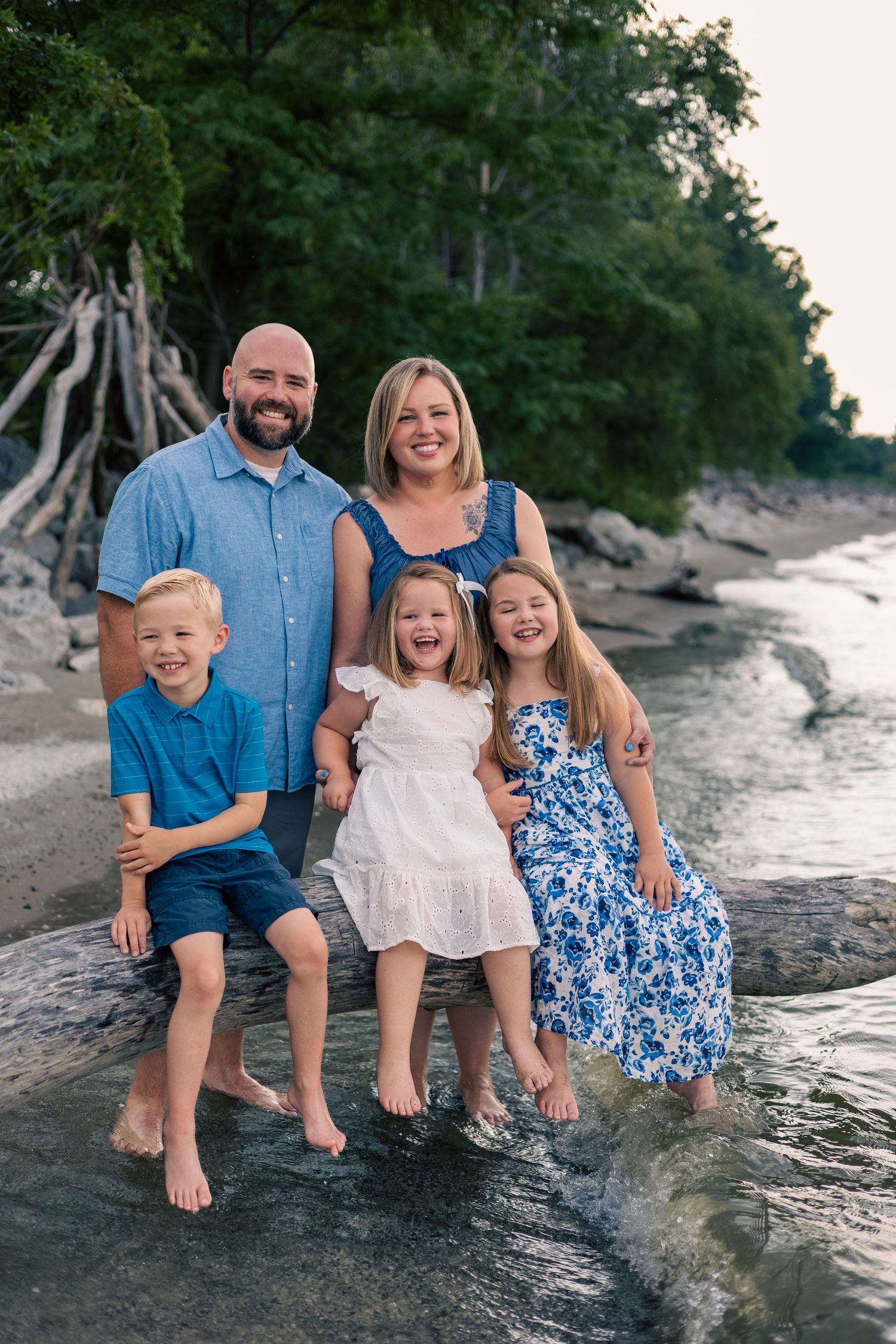 A family sits together on a rocky beach shoreline at sunset wearing coordinated blue and white outfits.