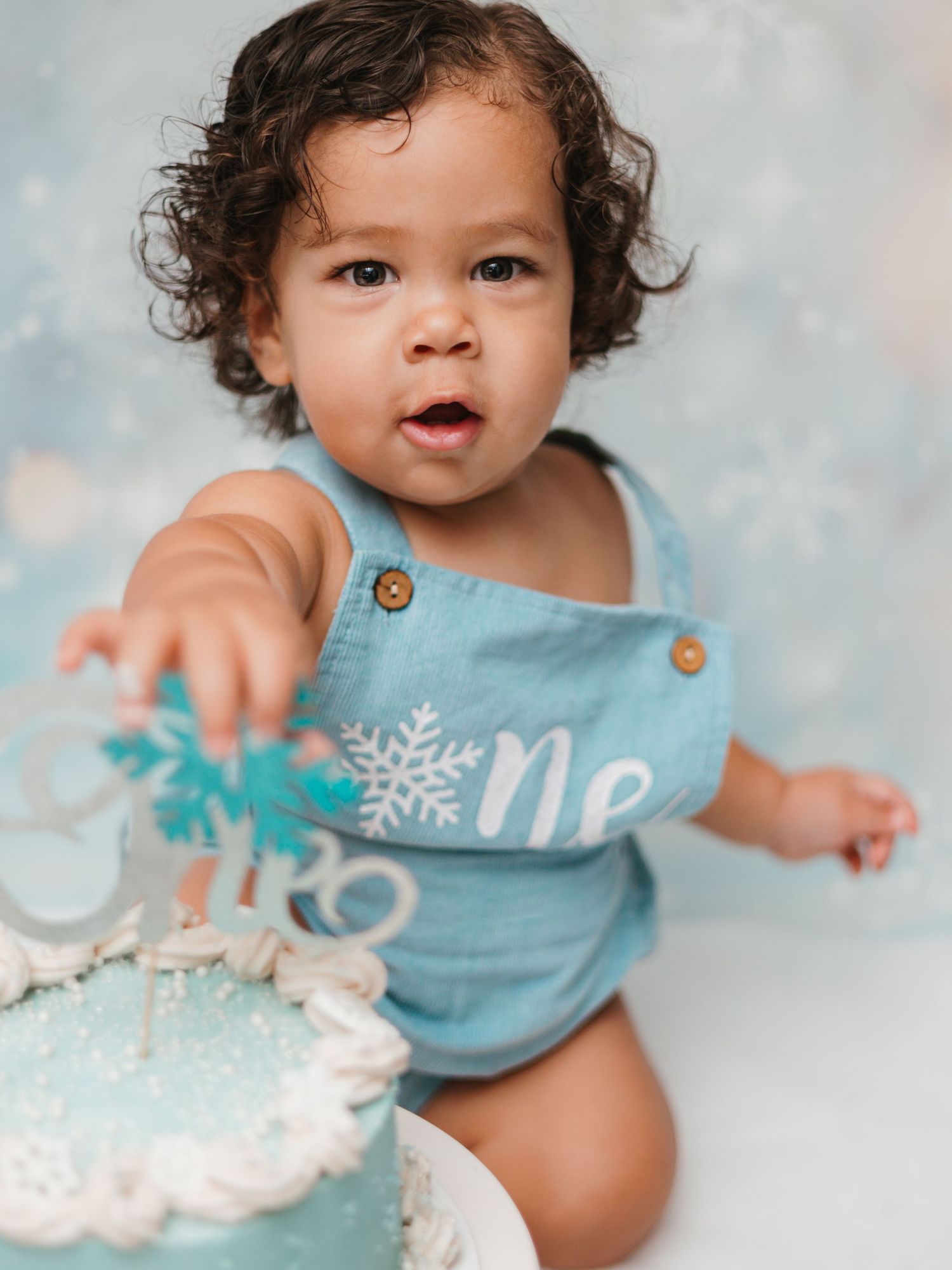 Baby in light blue romper with curly hair reaches toward a birthday cake with snowflake decorations.