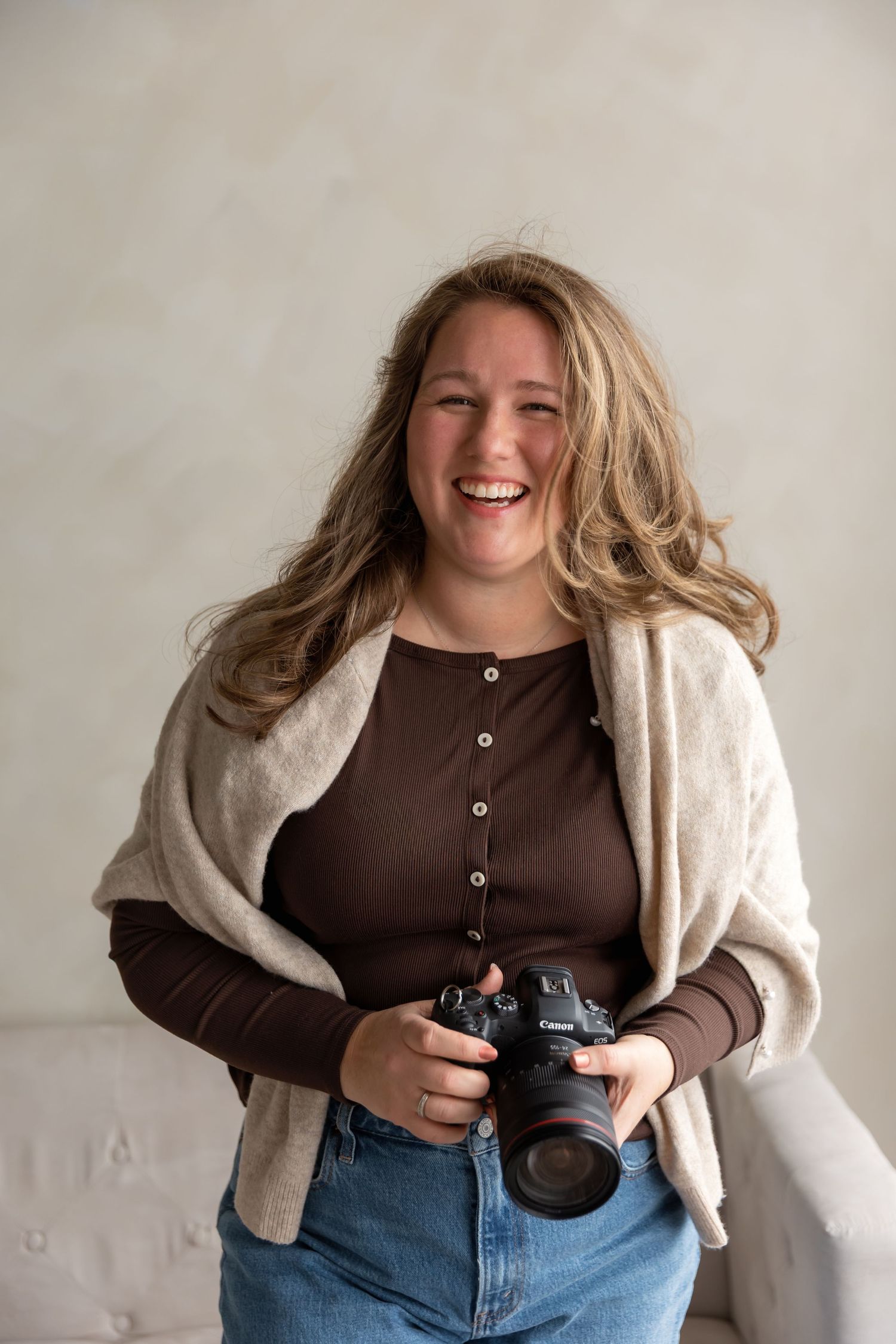 Smiling woman with long hair holds a DSLR camera, wearing brown top, jeans, and beige wrap in a bright studio.