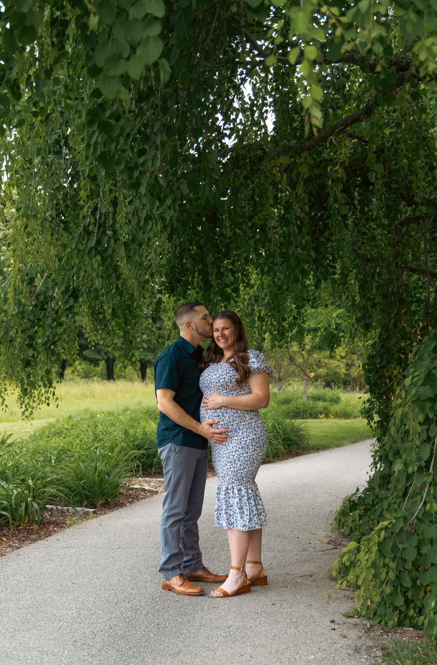Couple poses together on winding path beneath drooping willow branches in garden setting.