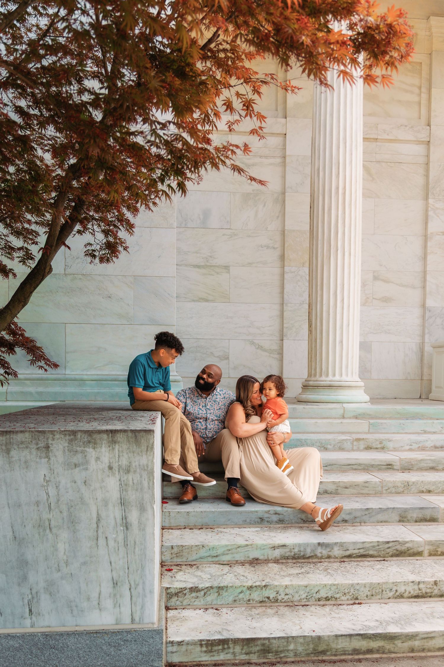 Family of four sitting on marble steps near a column and autumn tree at a historic monument.