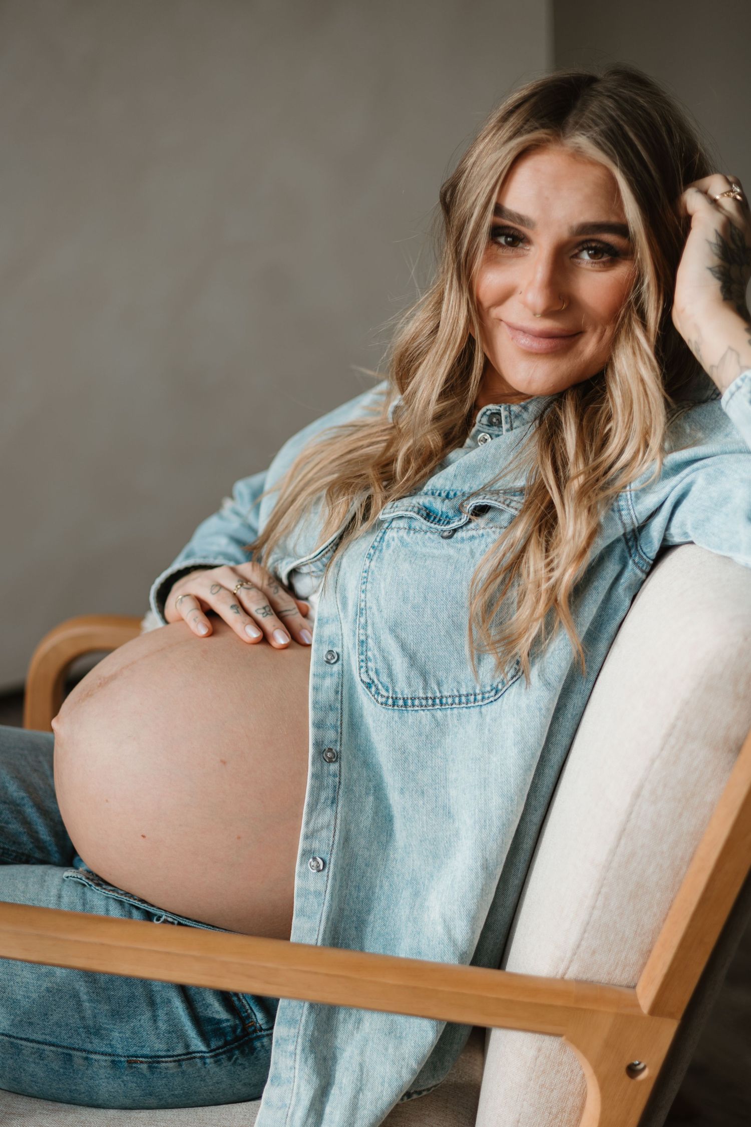 Woman in light denim jacket poses casually on beige couch during lifestyle photoshoot.