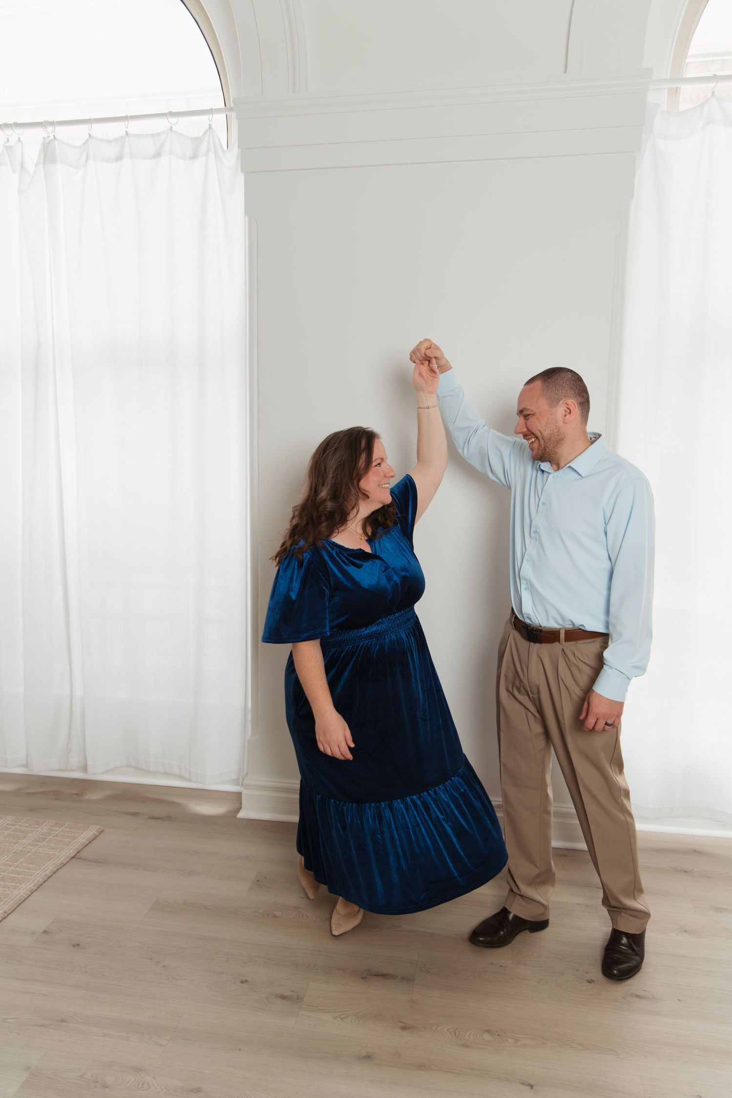 A couple dances together in a bright white room, with the woman wearing a navy blue dress and the man in light blue shirt.