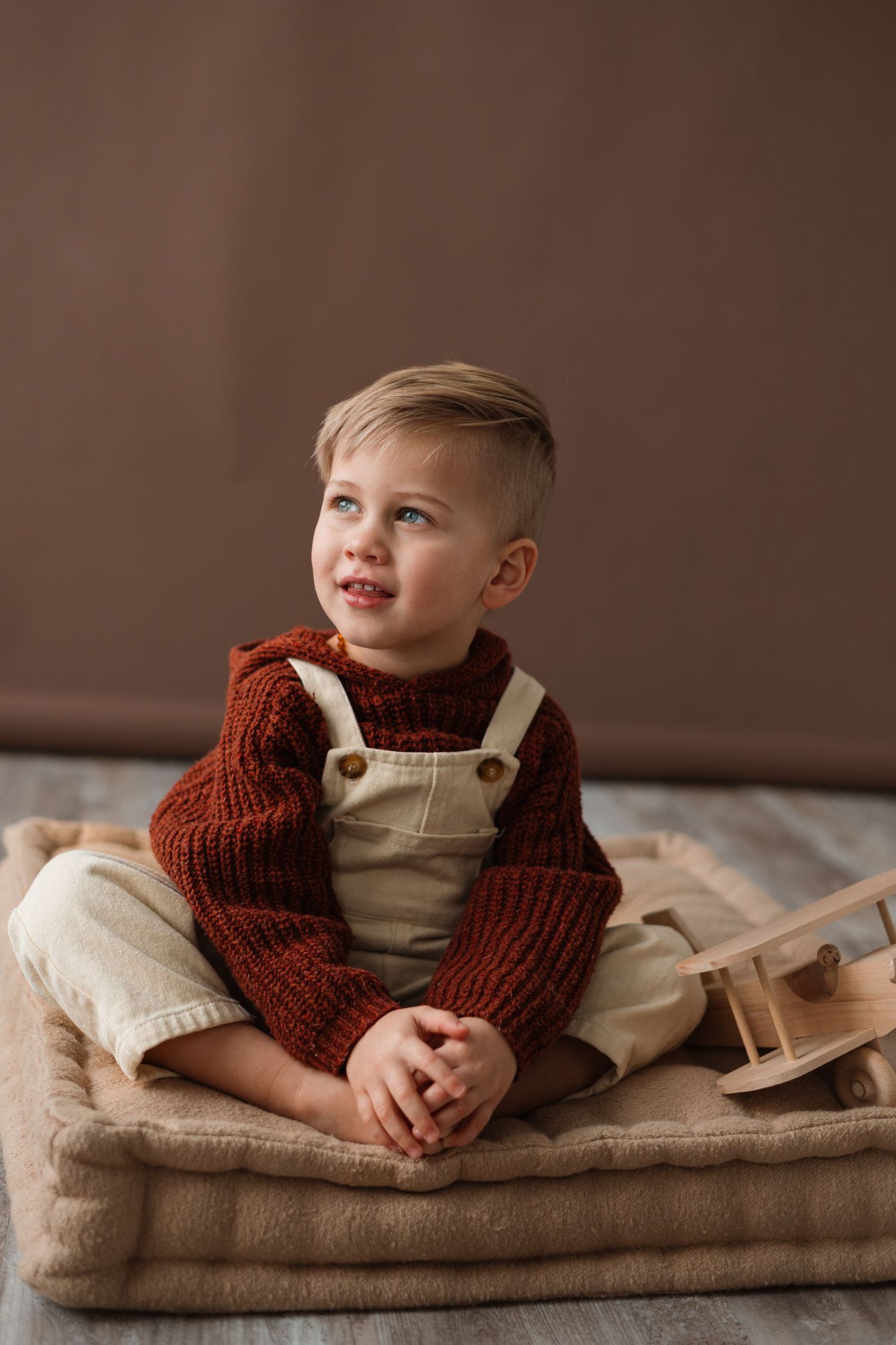 A boy in a brown knit sweater and cream overalls lies on a beige blanket in a warm studio setting.