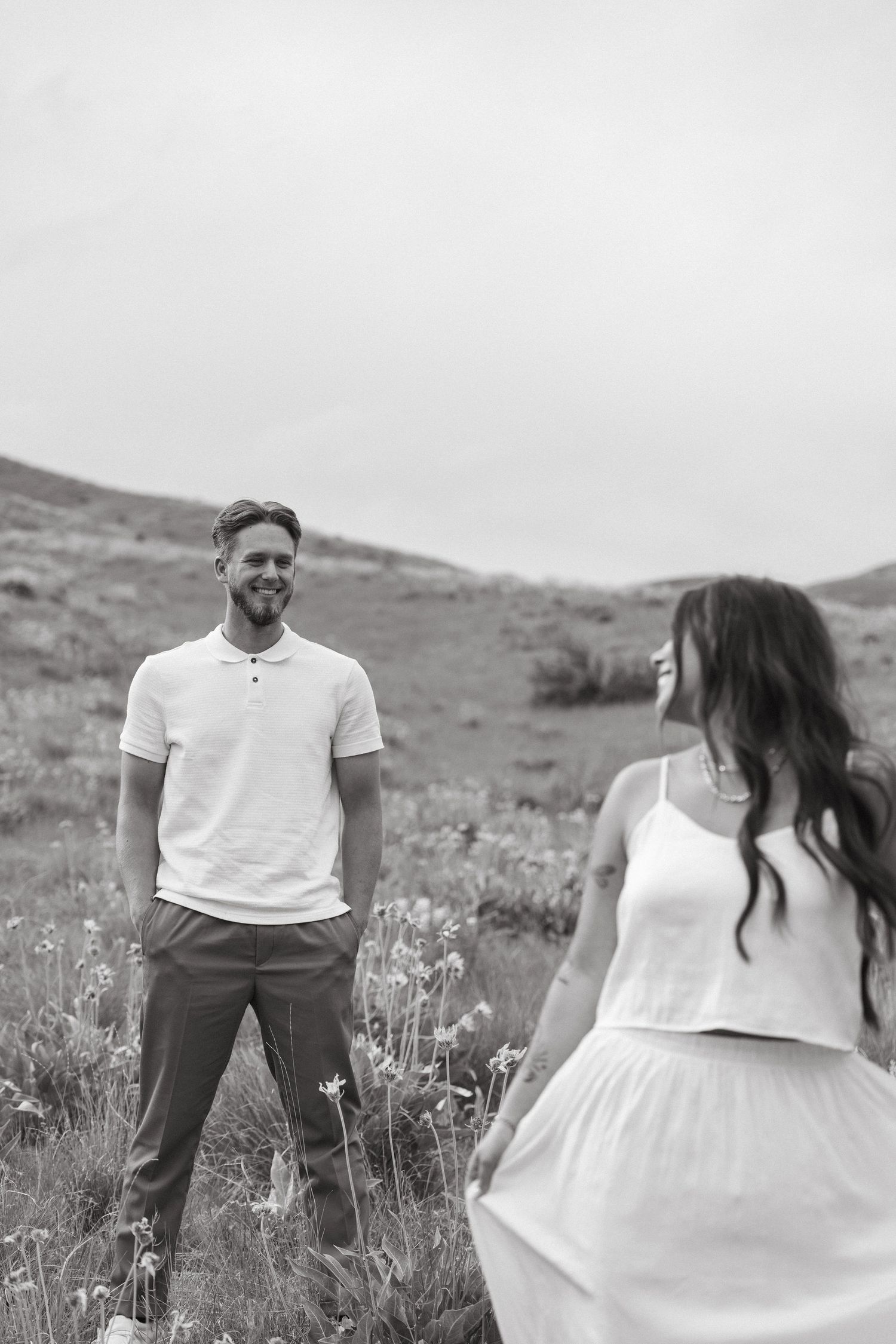 A couple in casual white outfits stand in a field of wildflowers on a hillside in black and white photography.