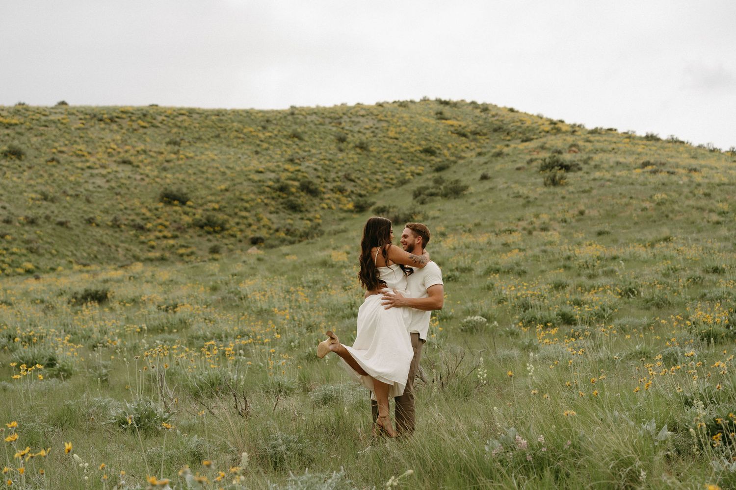 Couple embraces in flowing white dress against green hillside meadow on overcast day.