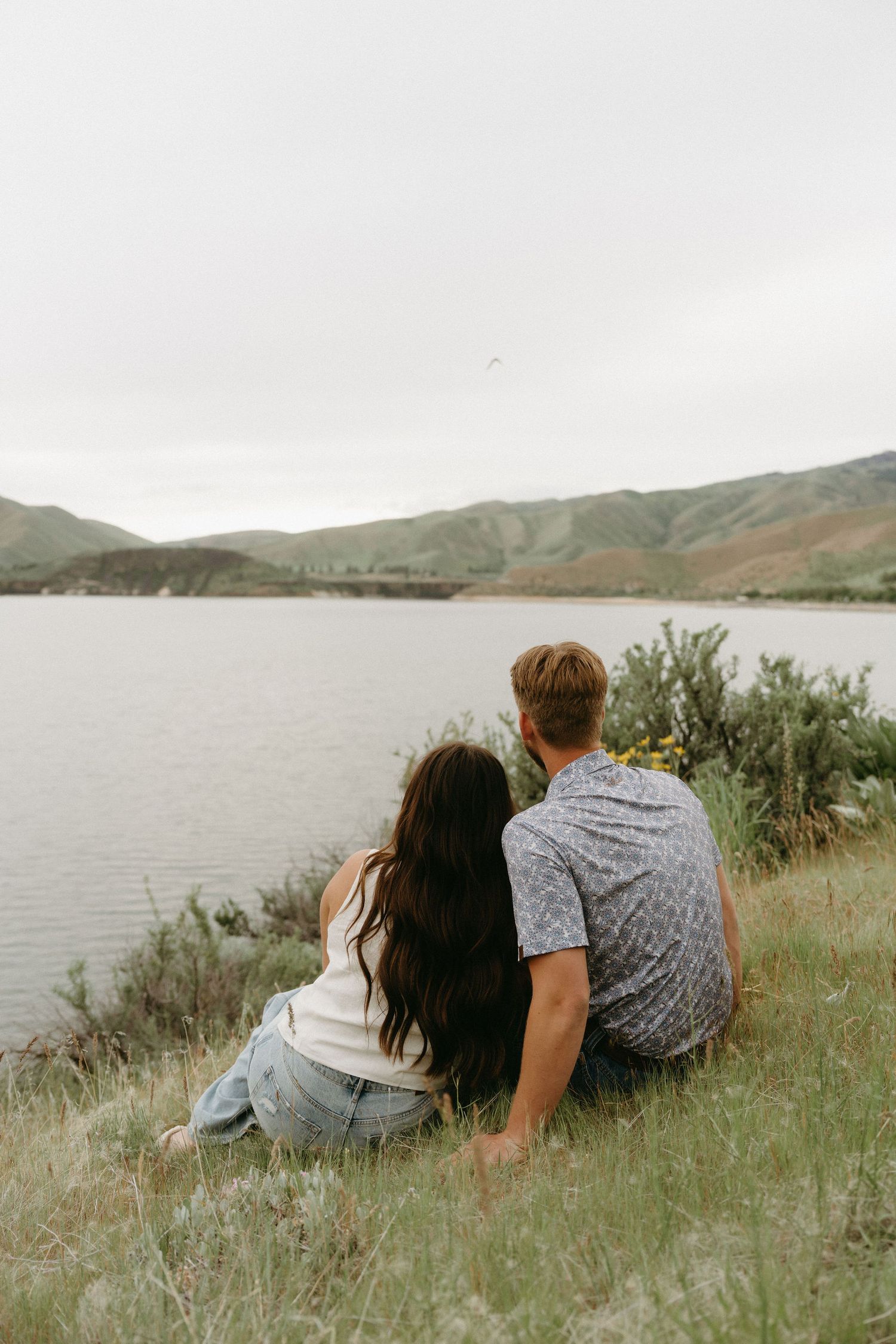 Couple lying together in tall grass by a serene lake with mountains in the background.