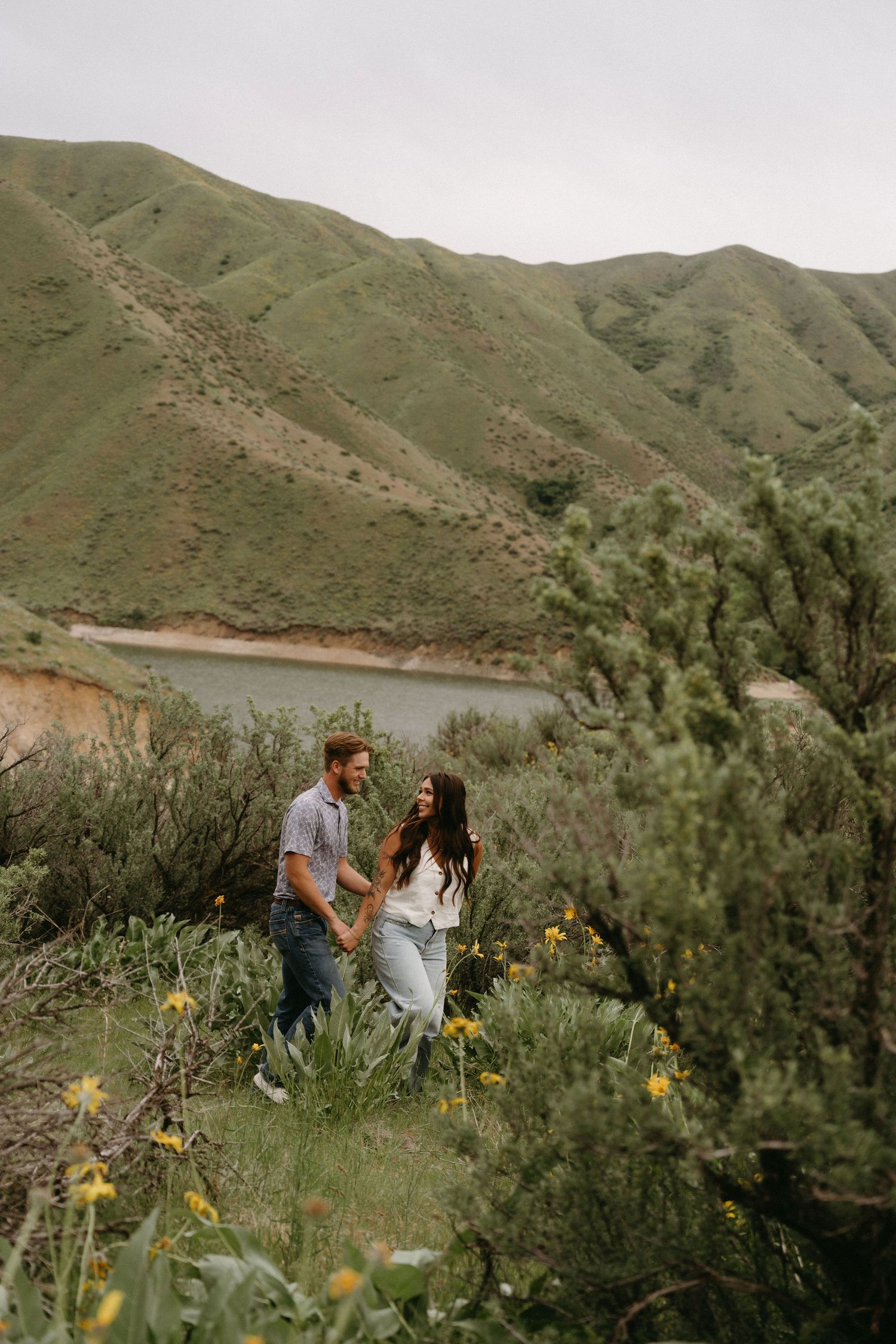 A couple stands together on a hillside overlooking a lake surrounded by green mountains and yellow wildflowers.