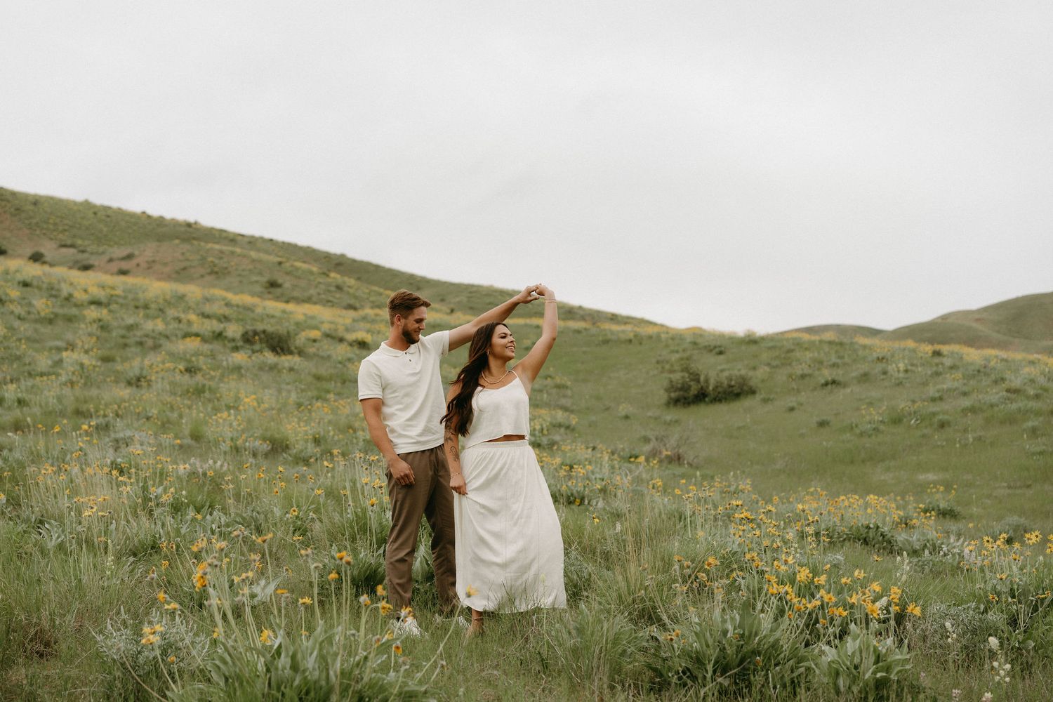 A couple dances together on a green hillside meadow with wildflowers under an overcast sky.