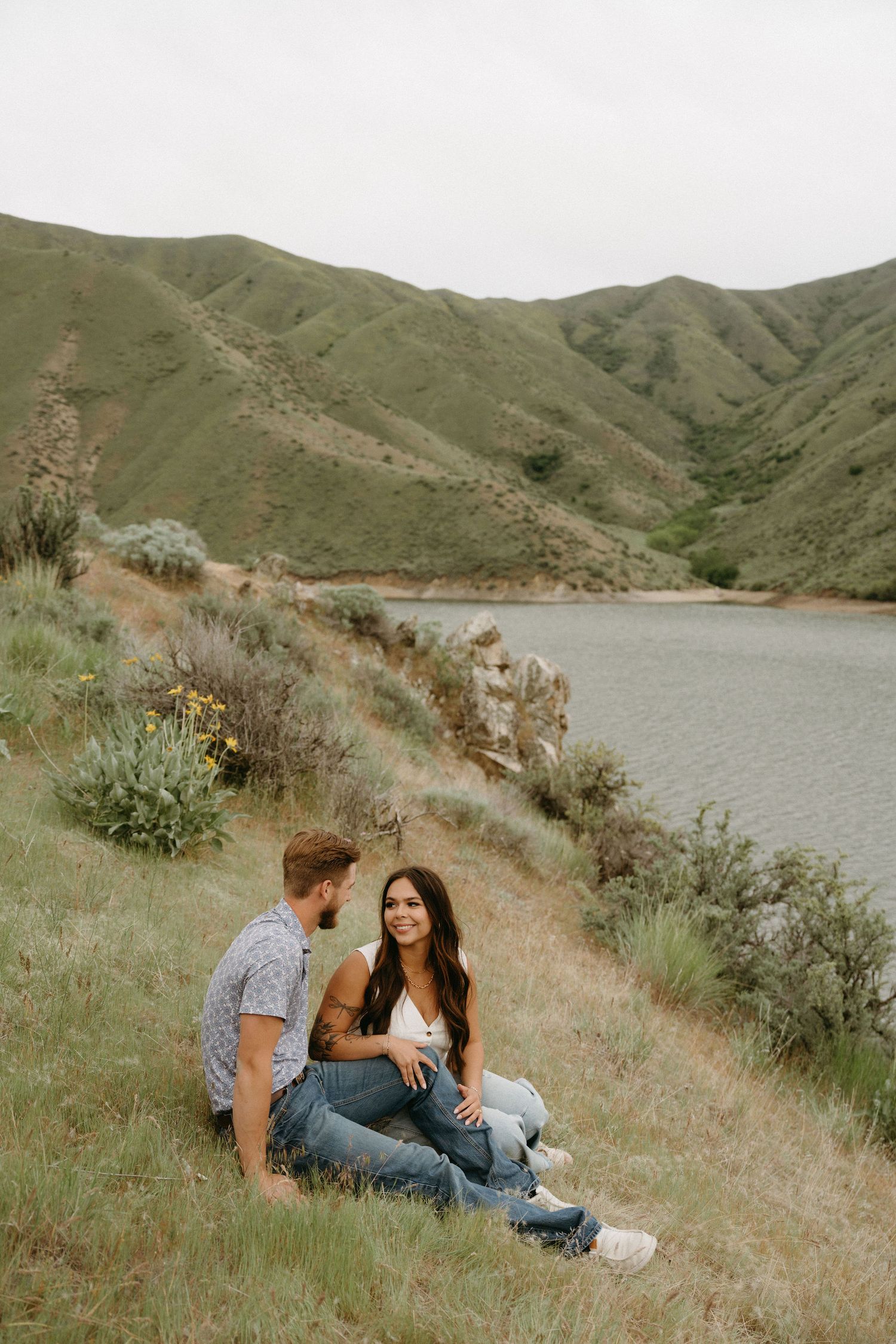 A couple sits together on a grassy hillside overlooking a serene lake surrounded by rolling green mountains.