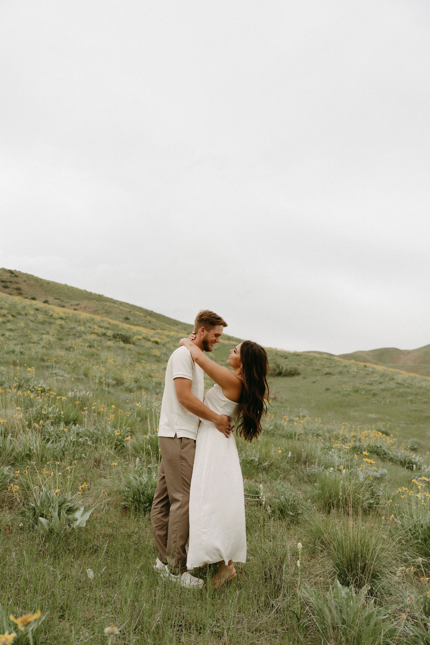 Couple embraces on a grassy hillside with wildflowers under an overcast sky in romantic outdoor setting.