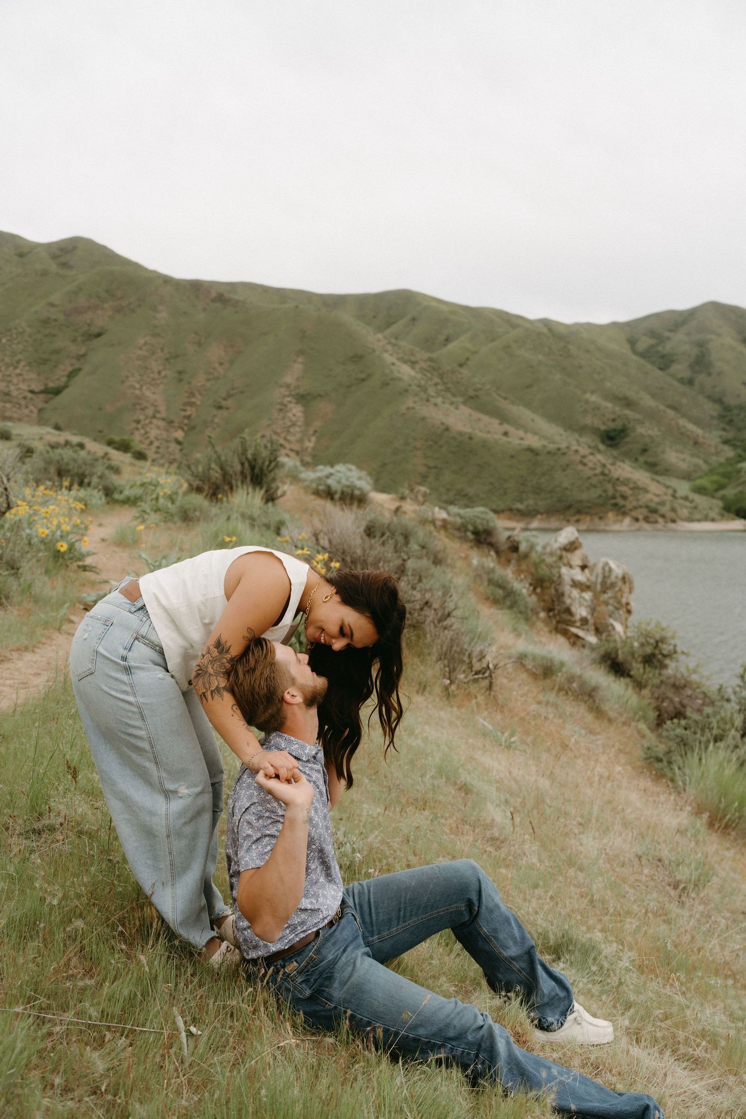Two people share an intimate moment on a grassy hillside overlooking a lake with scenic mountains in the background.