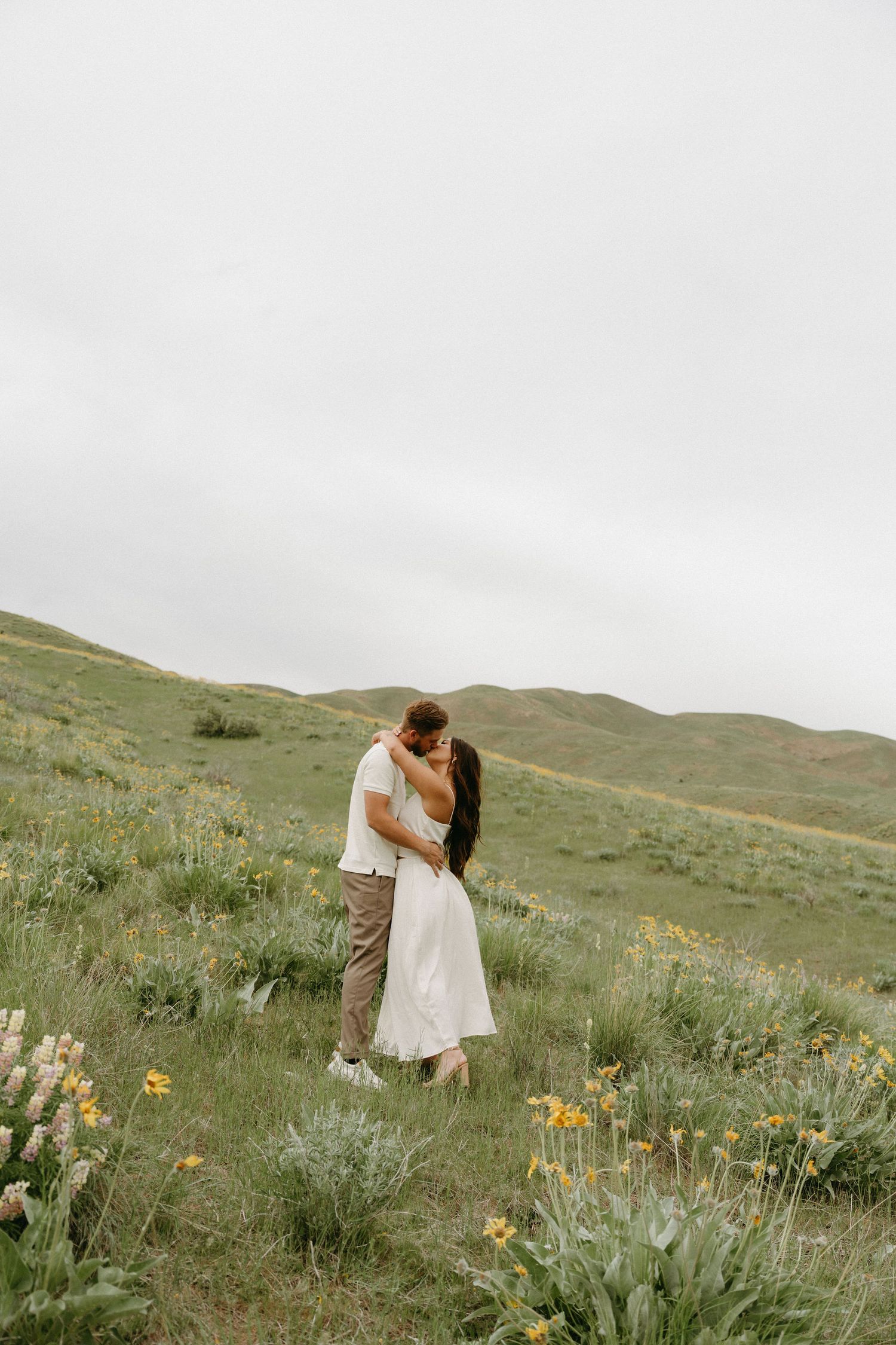Couple embraces romantically in a green hillside meadow filled with wildflowers on an overcast day.