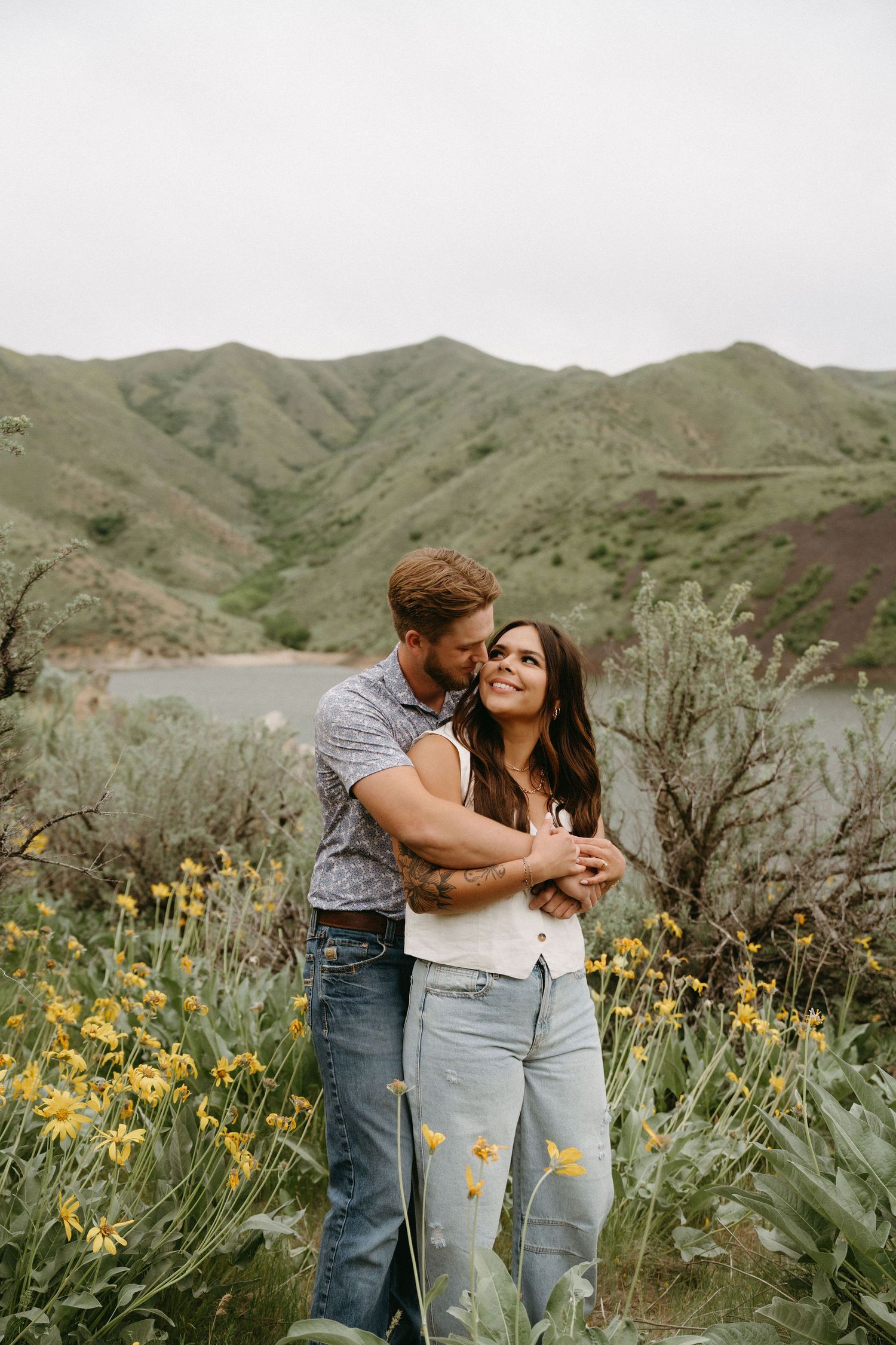 Romantic couple embraces while standing among yellow wildflowers in front of scenic mountain lake landscape.