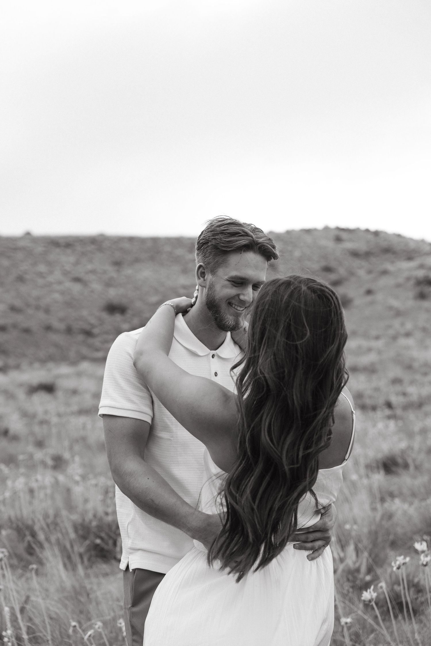A romantic black and white portrait of a couple embracing in a rustic outdoor setting with rolling hills in the background.