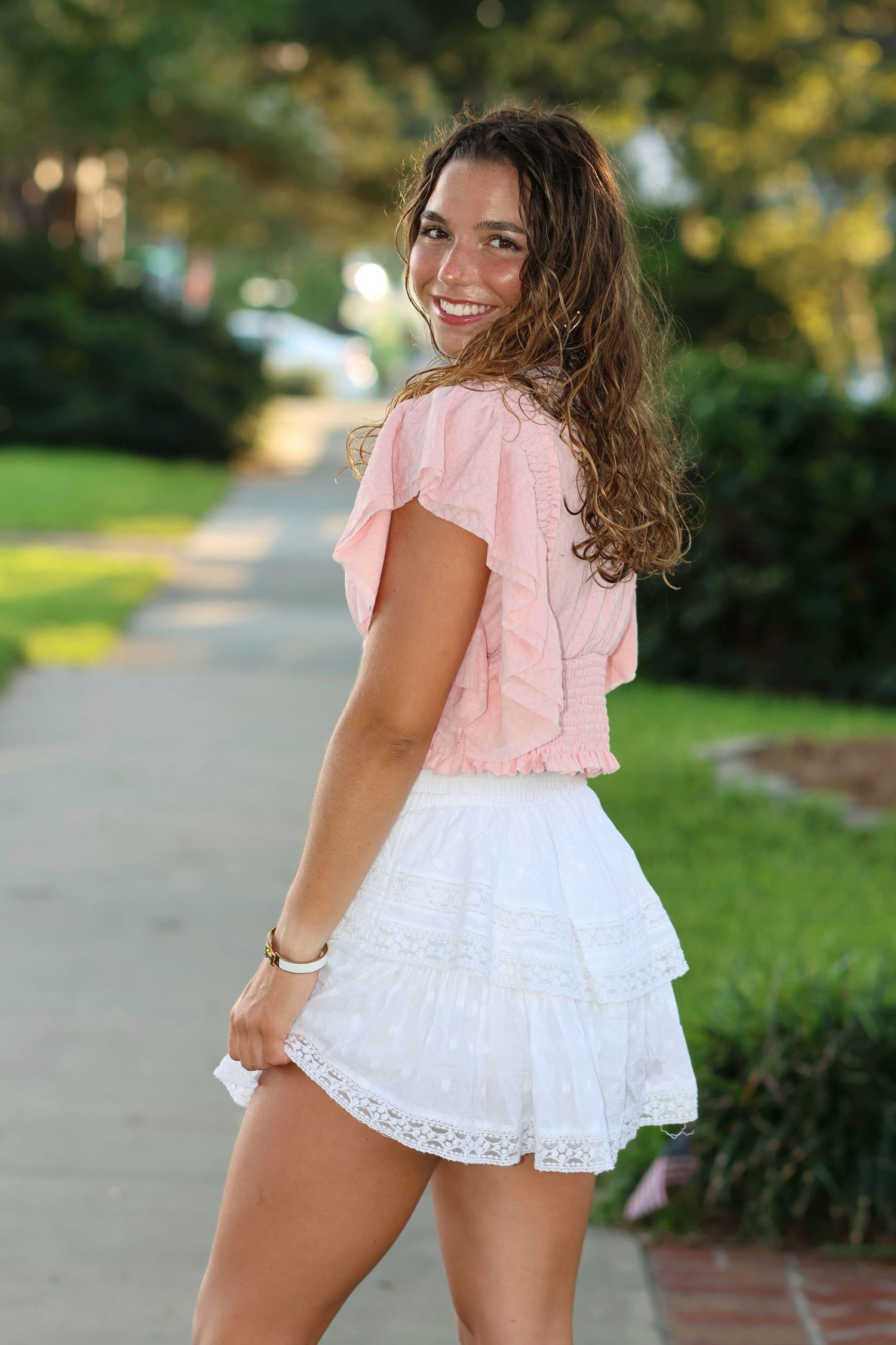 High school senior in a short skirt with lace and a pale pink top looking over her shoulder smiling walking down a tree lined st. at sunset.