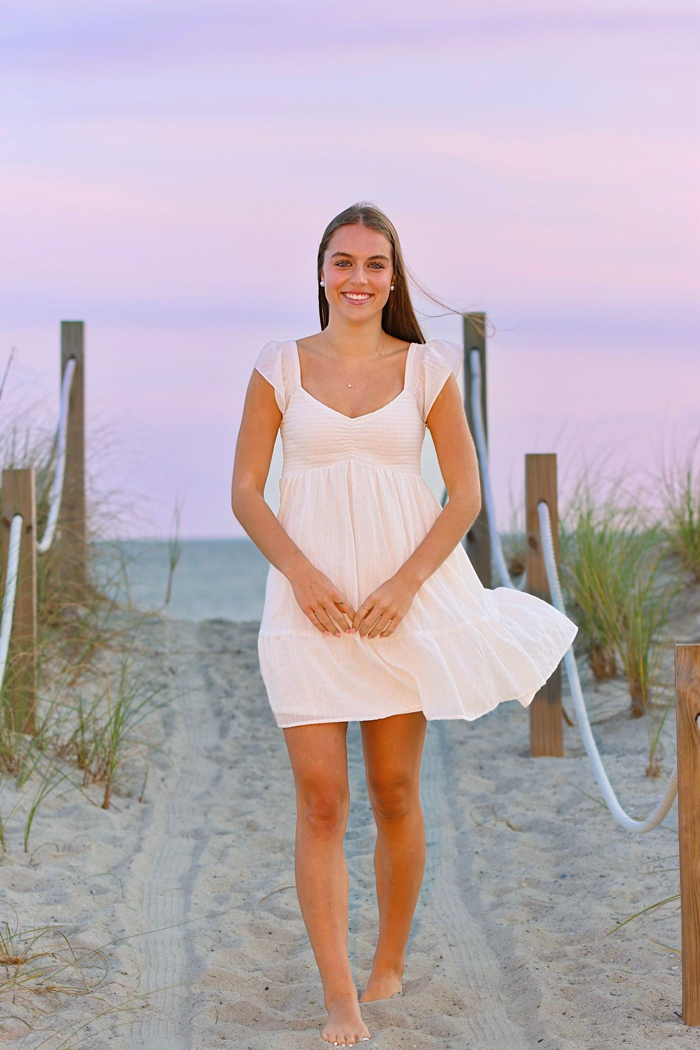 High school senior girl on the beach in a short white dress at sunset walking down a path in the sand.
