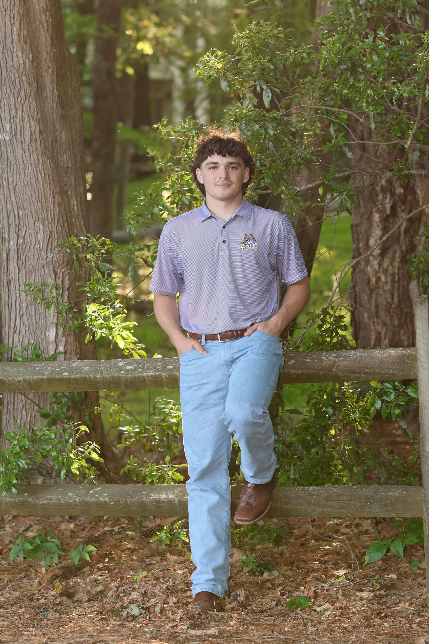 High school senior guy standing leaning against a fence with hands in pockets wearing a polo shirt and jeans.