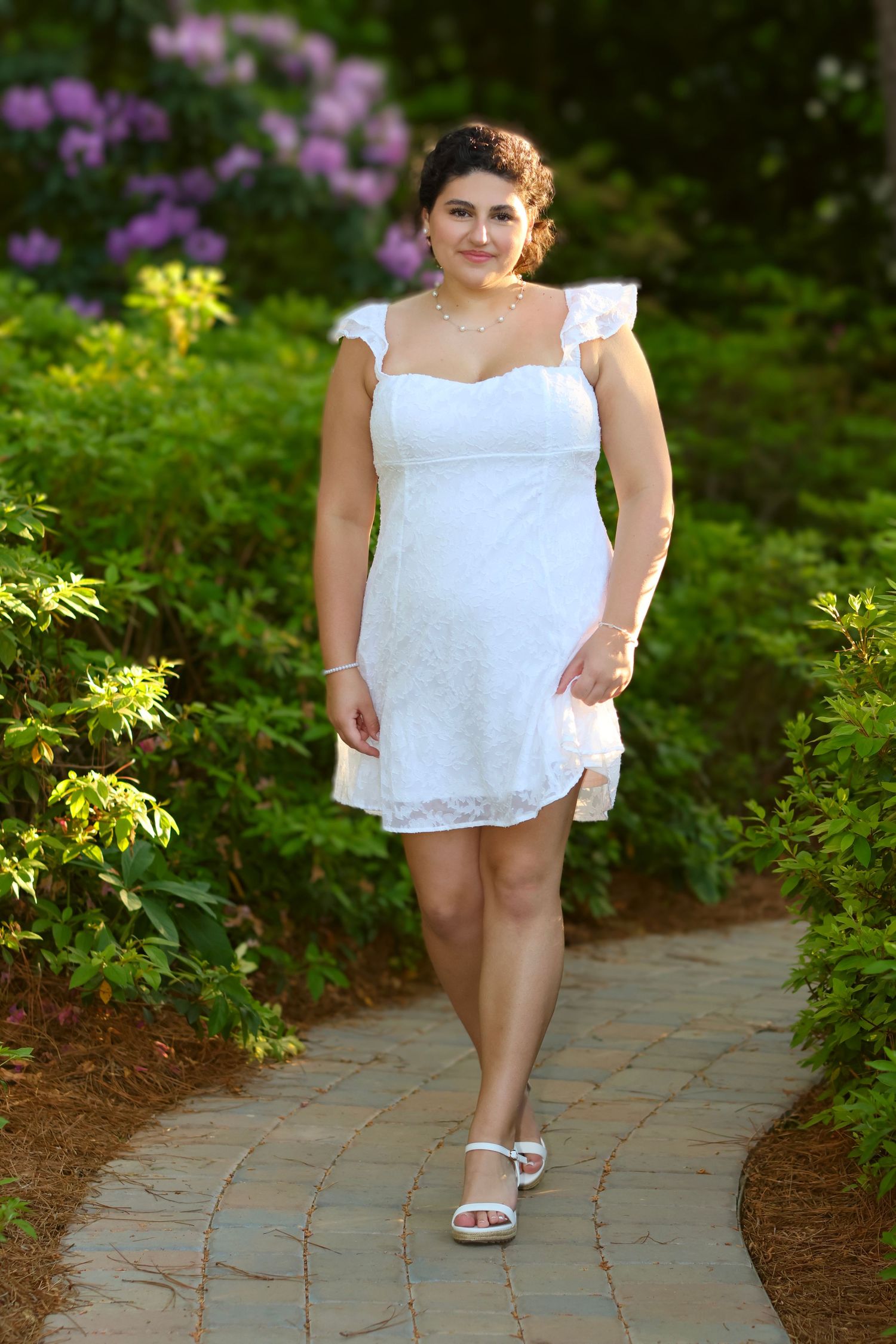 High school senior girl in a beautiful white dress walking confidently in a garden at sunset.