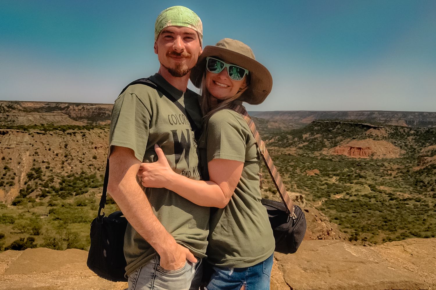 Two hikers in matching green shirts embrace while sightseeing at a scenic desert canyon overlook.