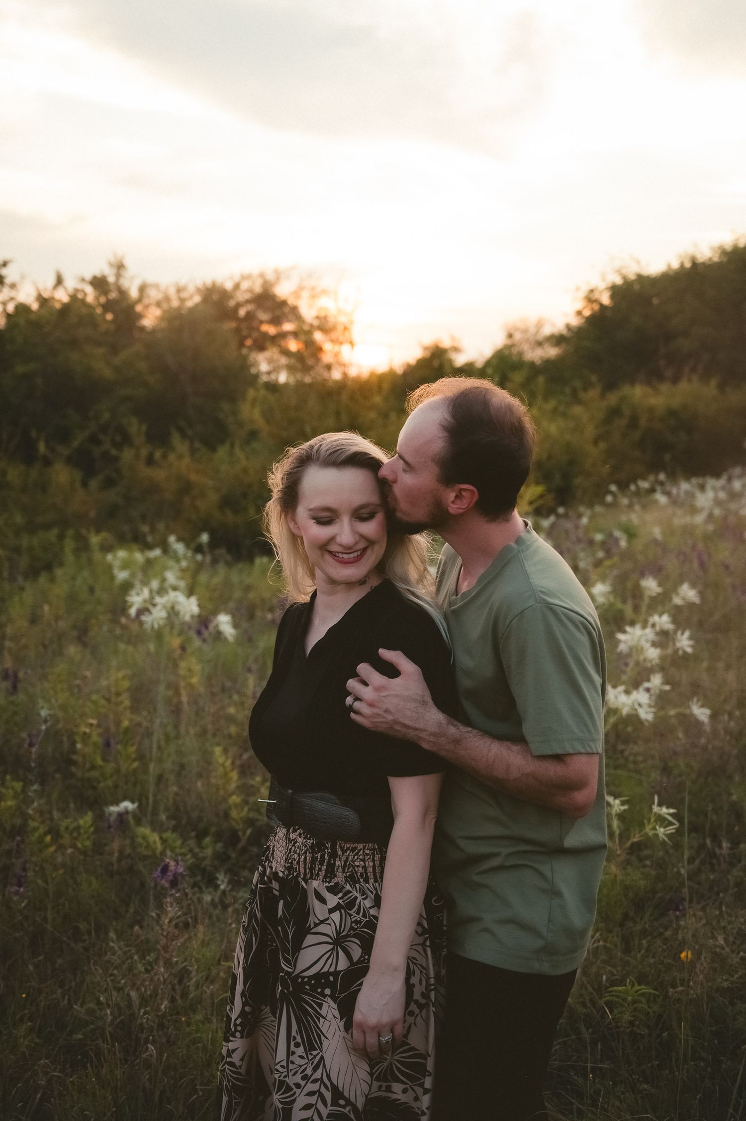 A couple shares an intimate moment in a wildflower meadow at sunset, surrounded by tall grass and trees.