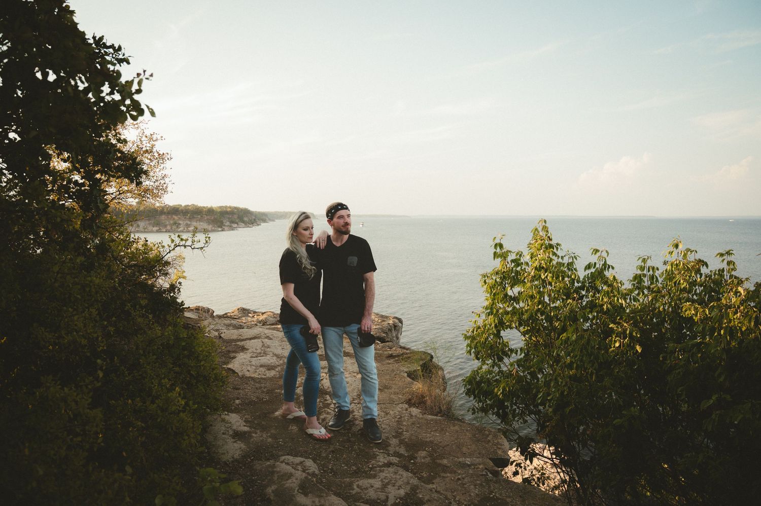 A couple stands together on a rocky coastal cliff overlooking the ocean at sunset.